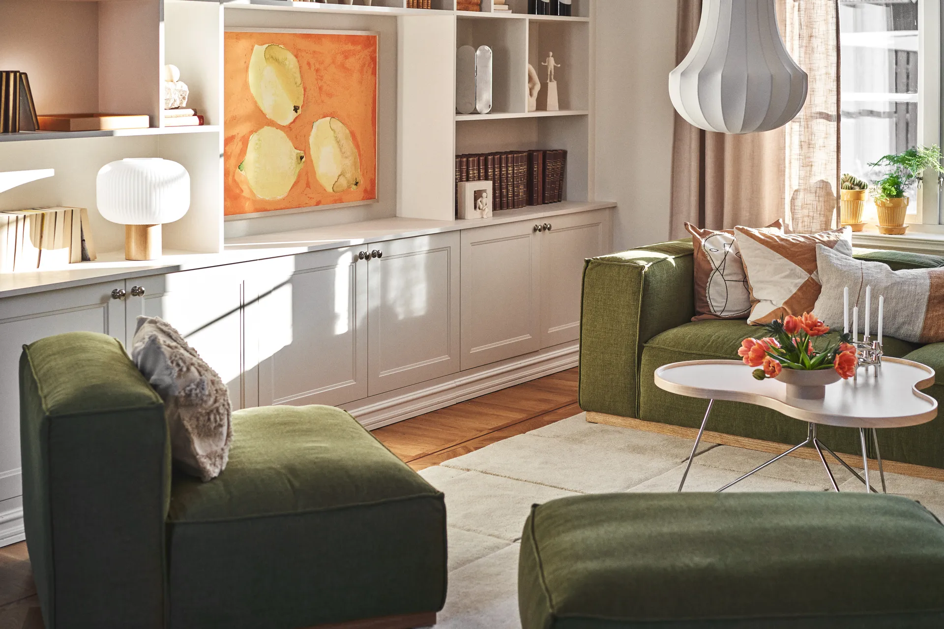 Solljust vardagsrum med gröna soffor, ett vitt soffbord med orange tulpaner och ljus, samt vita inbyggda hyllor med konst.