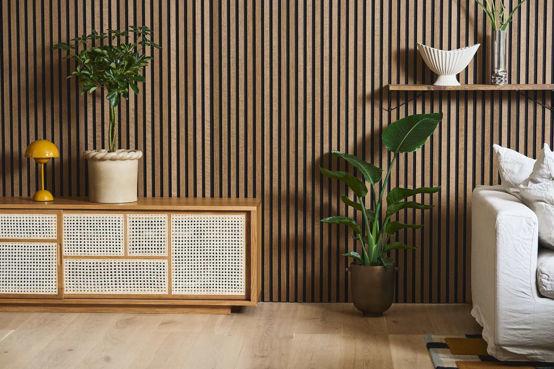 Ett modernt vardagsrum med en ribbad trävägg, rottingkabinett, gul lampa, krukväxter och en vit soffa.