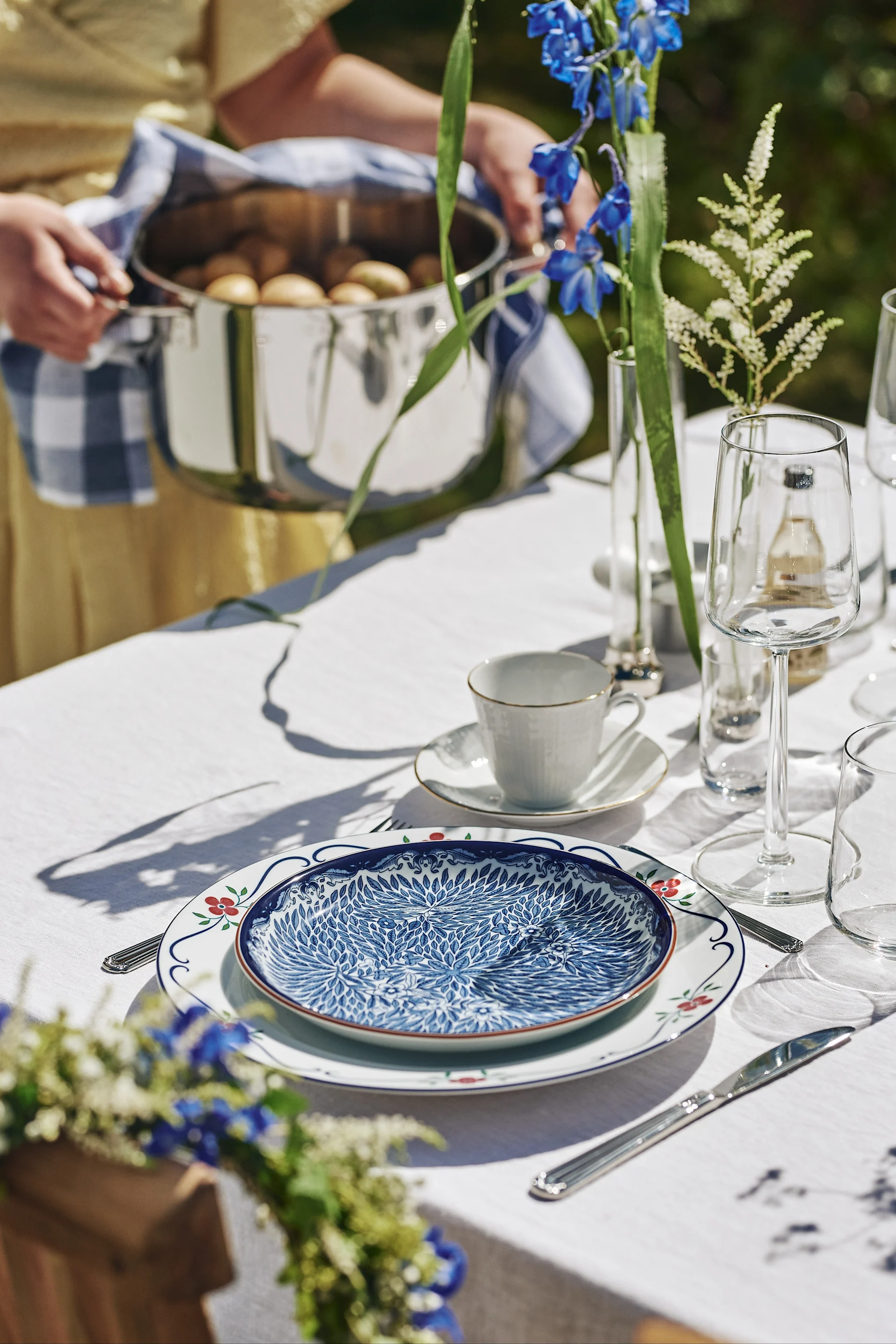 Midsommarkrans med blåa och vita blommor hängandes på stol vid den blåvita midsommardukningen.