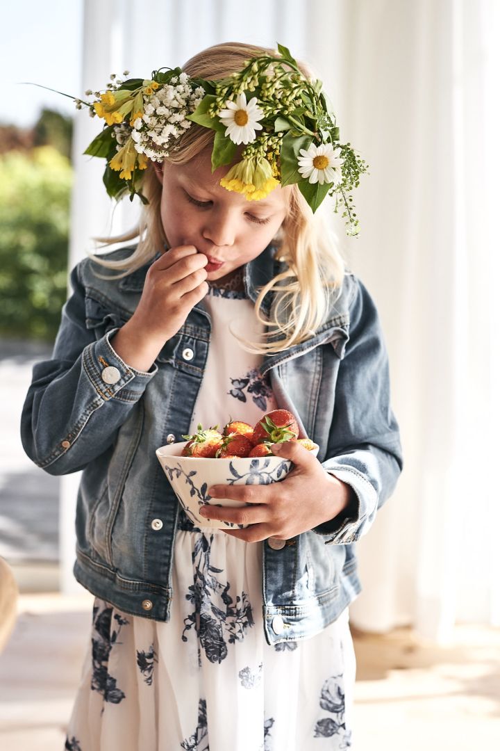 Flicka med midsommarkrans i håret äter jordgubbar ur en blåvit skål från Scandi Living.