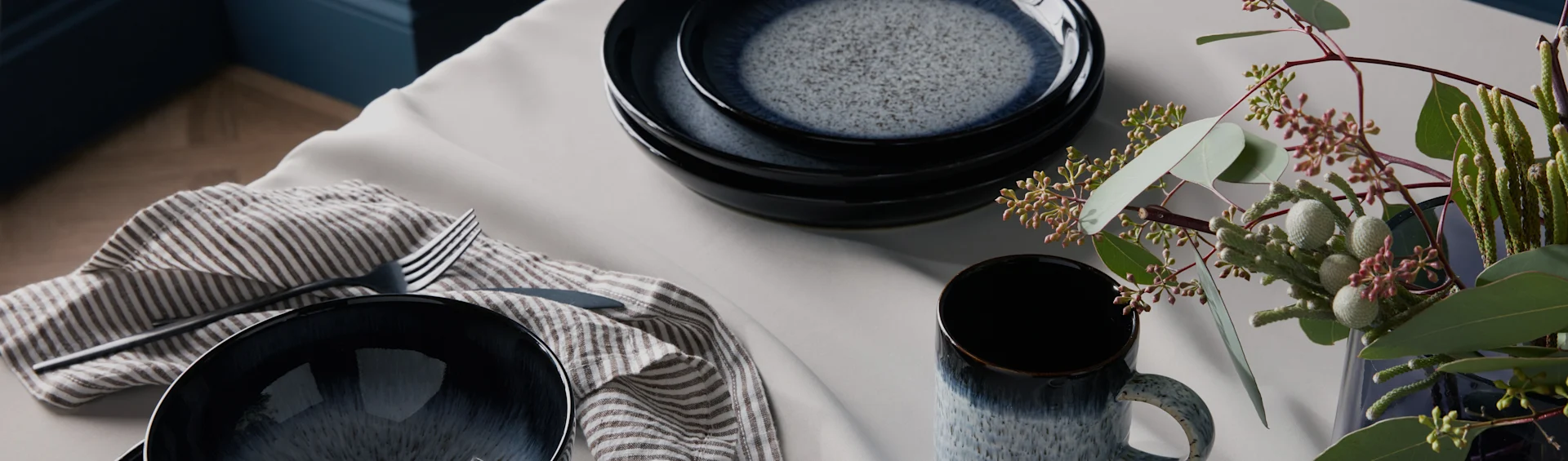 Spräckligt mörkblått keramikservis, randig servett, bestick och grönt bladverk på ett ljust bord.