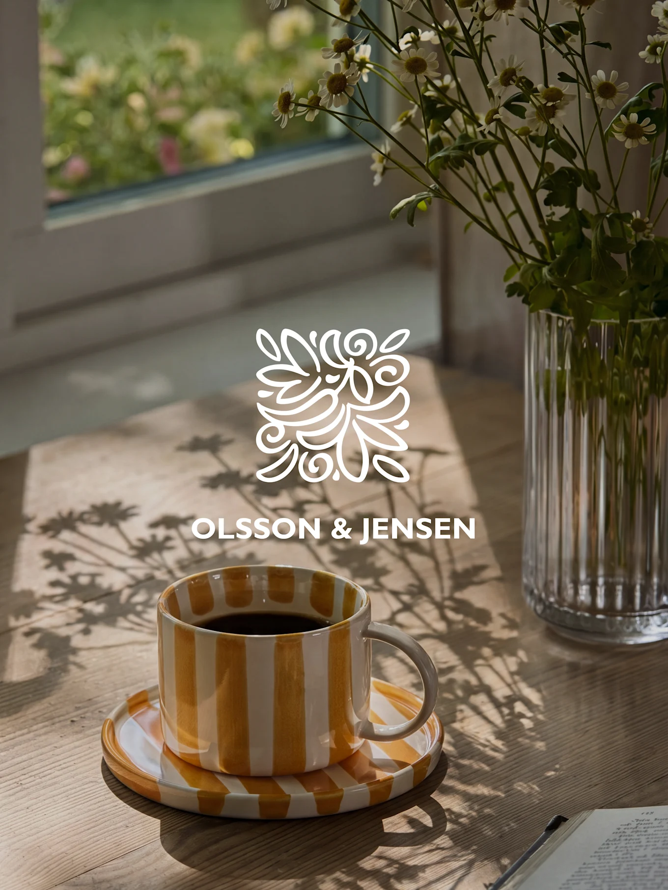 Gul- och vitrandig kaffemugg på ett träbord med en vas vita blommor och solbelysta skuggor, Olsson & Jensen-logotyp.