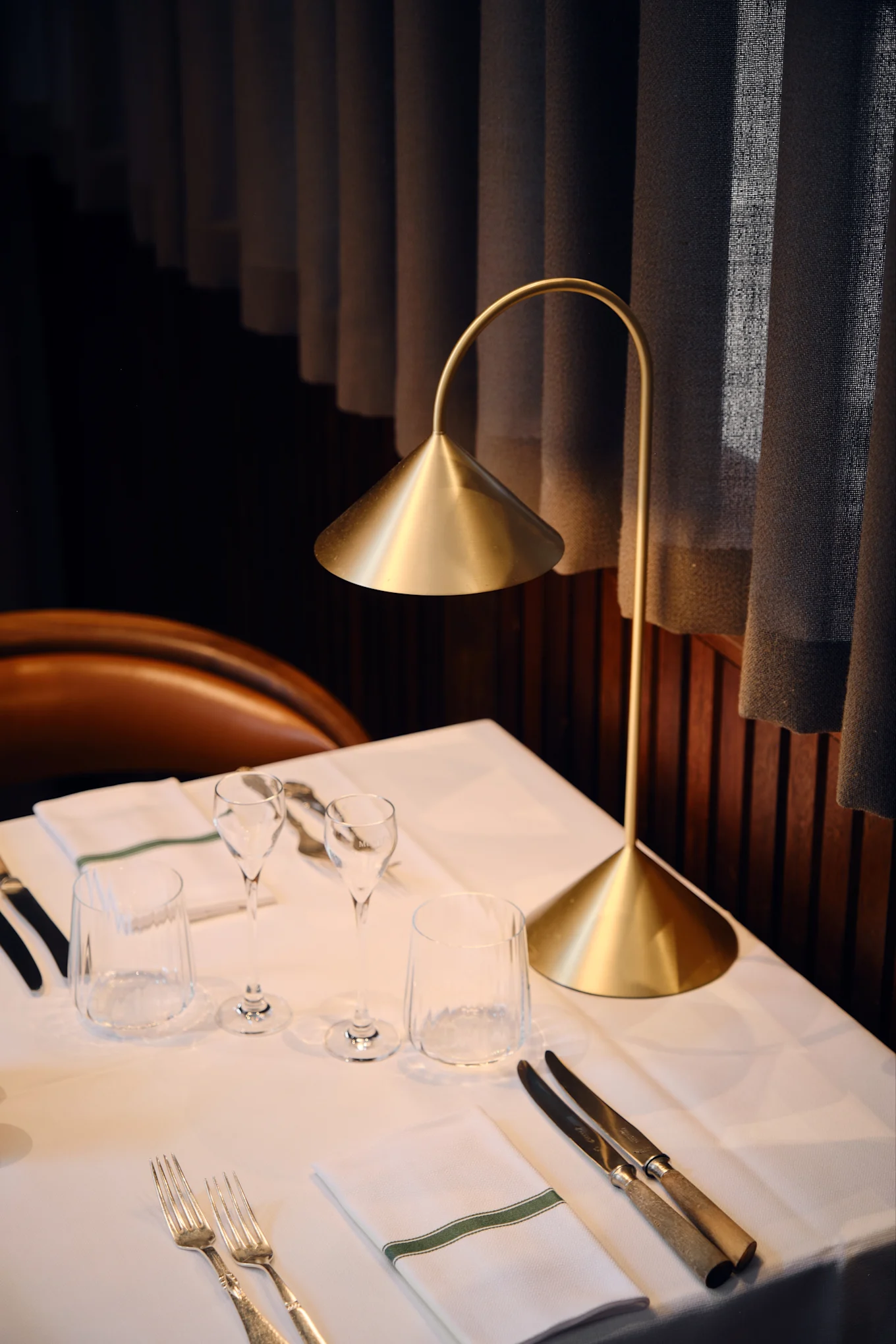 En modern guldbordslampa lyser upp ett dukat restaurangbord med vita dukar, glas, bestick och en servett med grön rand.