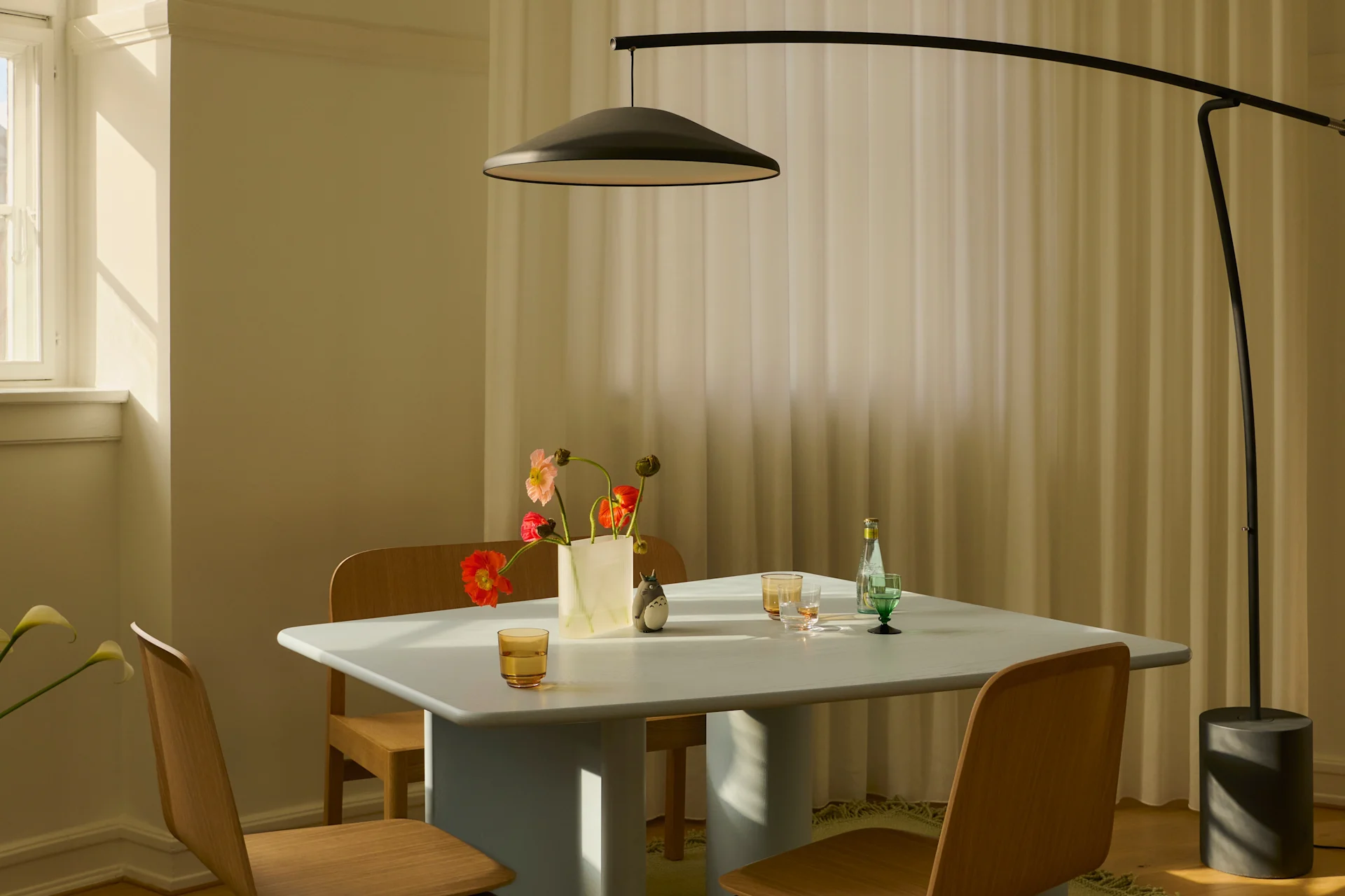 Modern matsal med ljusblått bord, trästolar, svart golvlampa och vas med blommor i solljus.