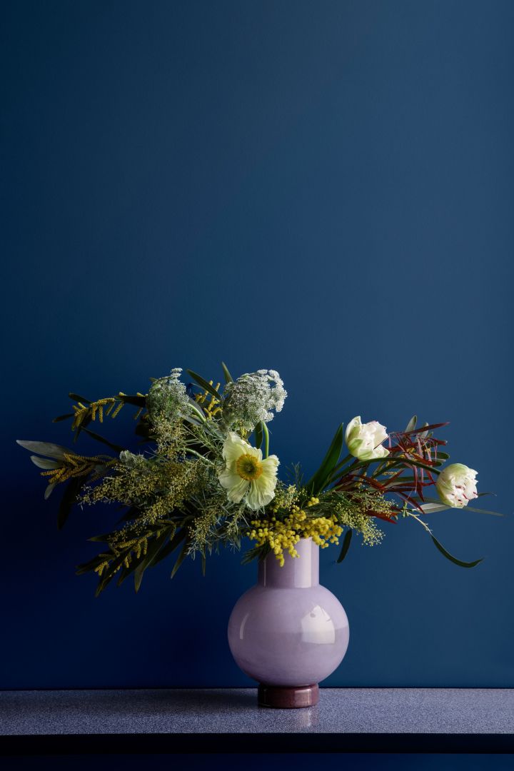 Lila, rund vas från Broste Copenhagen med vårblommor i mot en blå vägg - två färger vi gärna inreder med enligt färgtrenderna 2022.