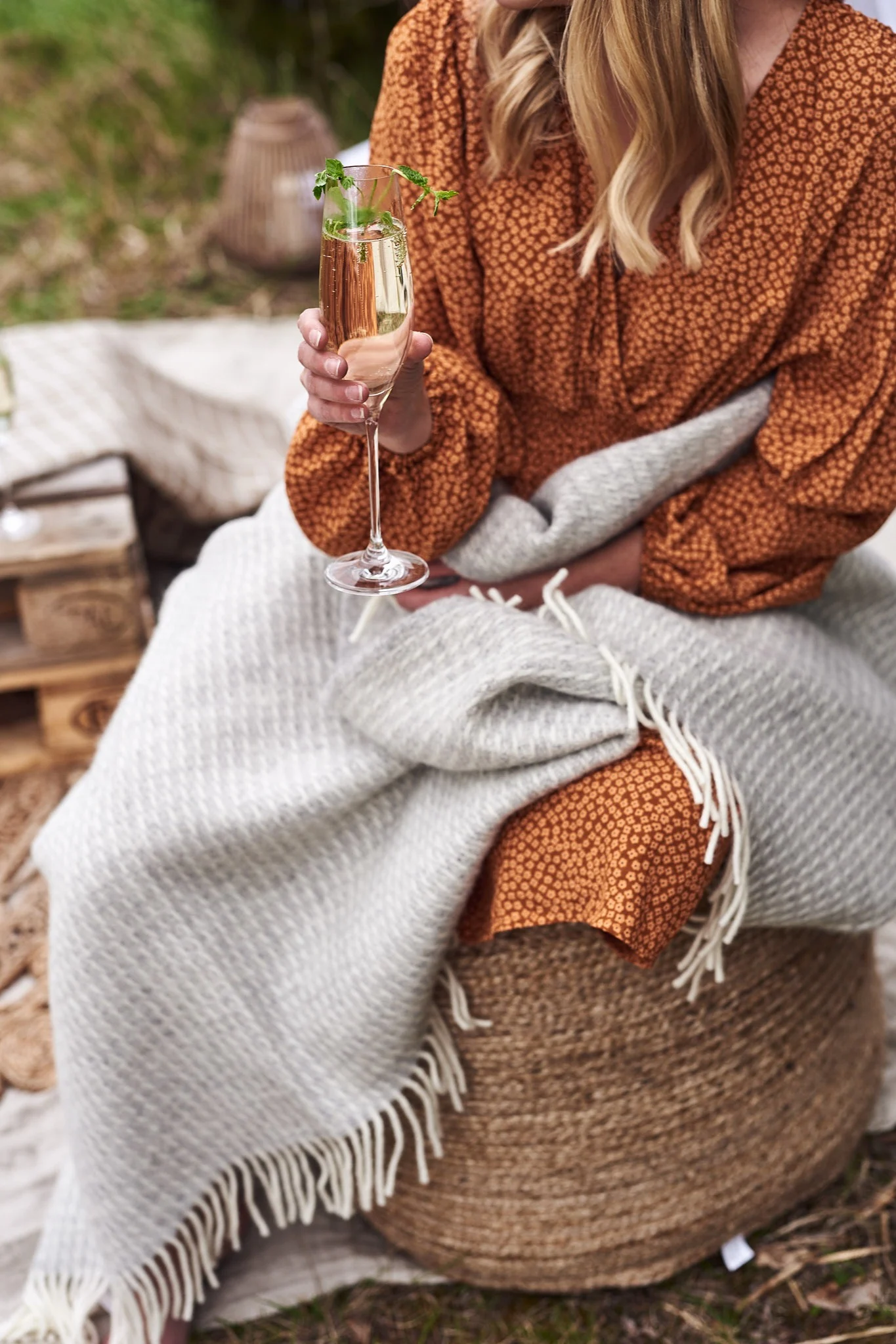 Tjej sitter på en sittpuff i jute med pläd över sig och ett glas bubbel i handen på en glamping-utflykt. 