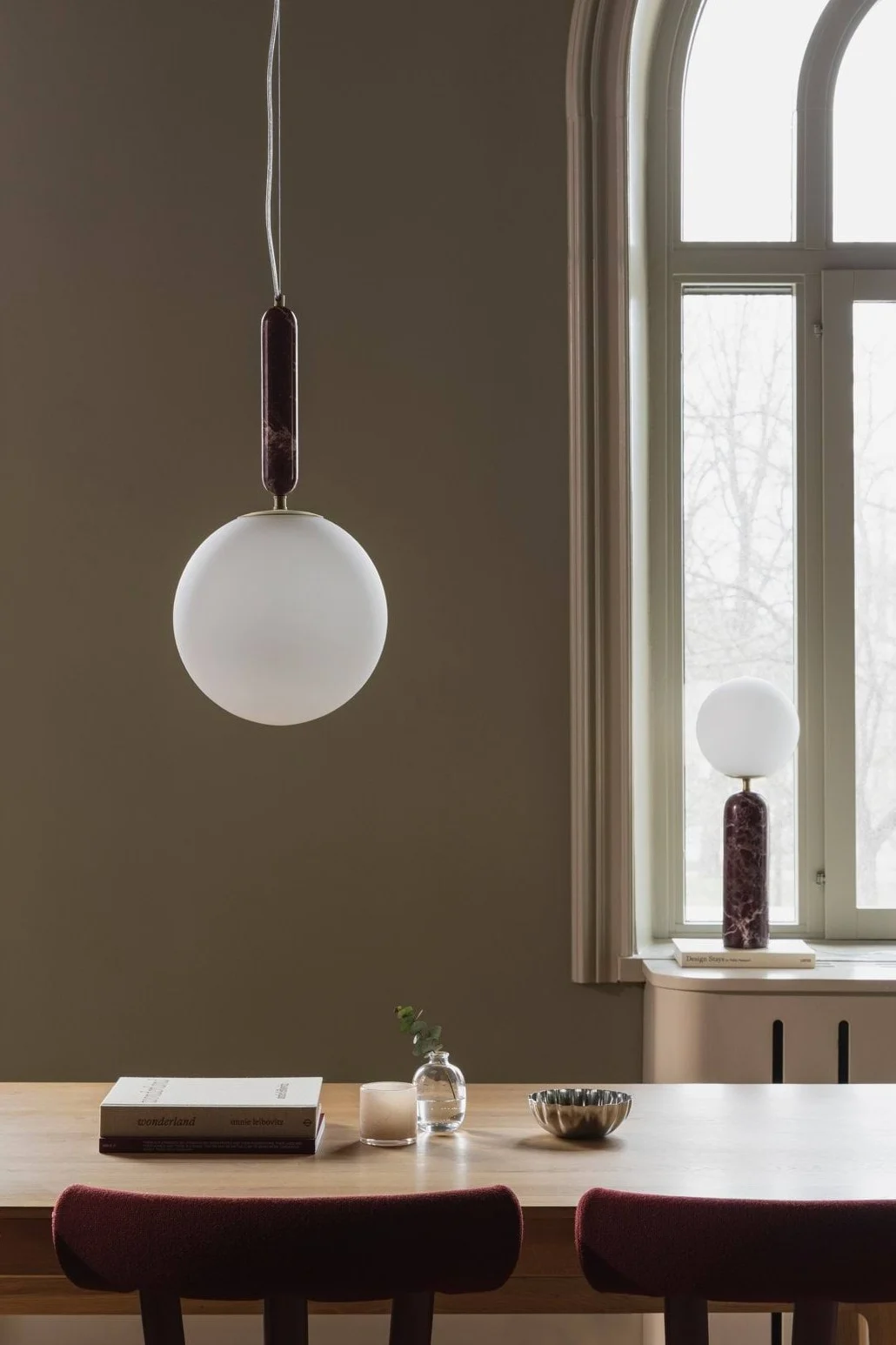 En matsal med träbord och vinröda stolar, med en sfärisk pendellampa och en matchande bordslampa med mörkröda marmordetaljer.