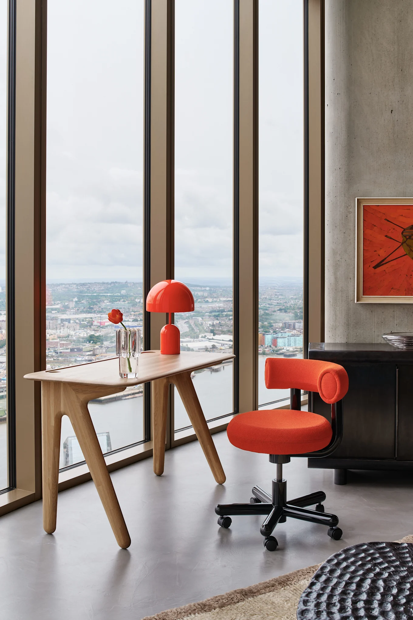 Ett modernt kontor med en orange snurrstol och matchande lampa vid ett ljust träskrivbord med utsikt över en stad.