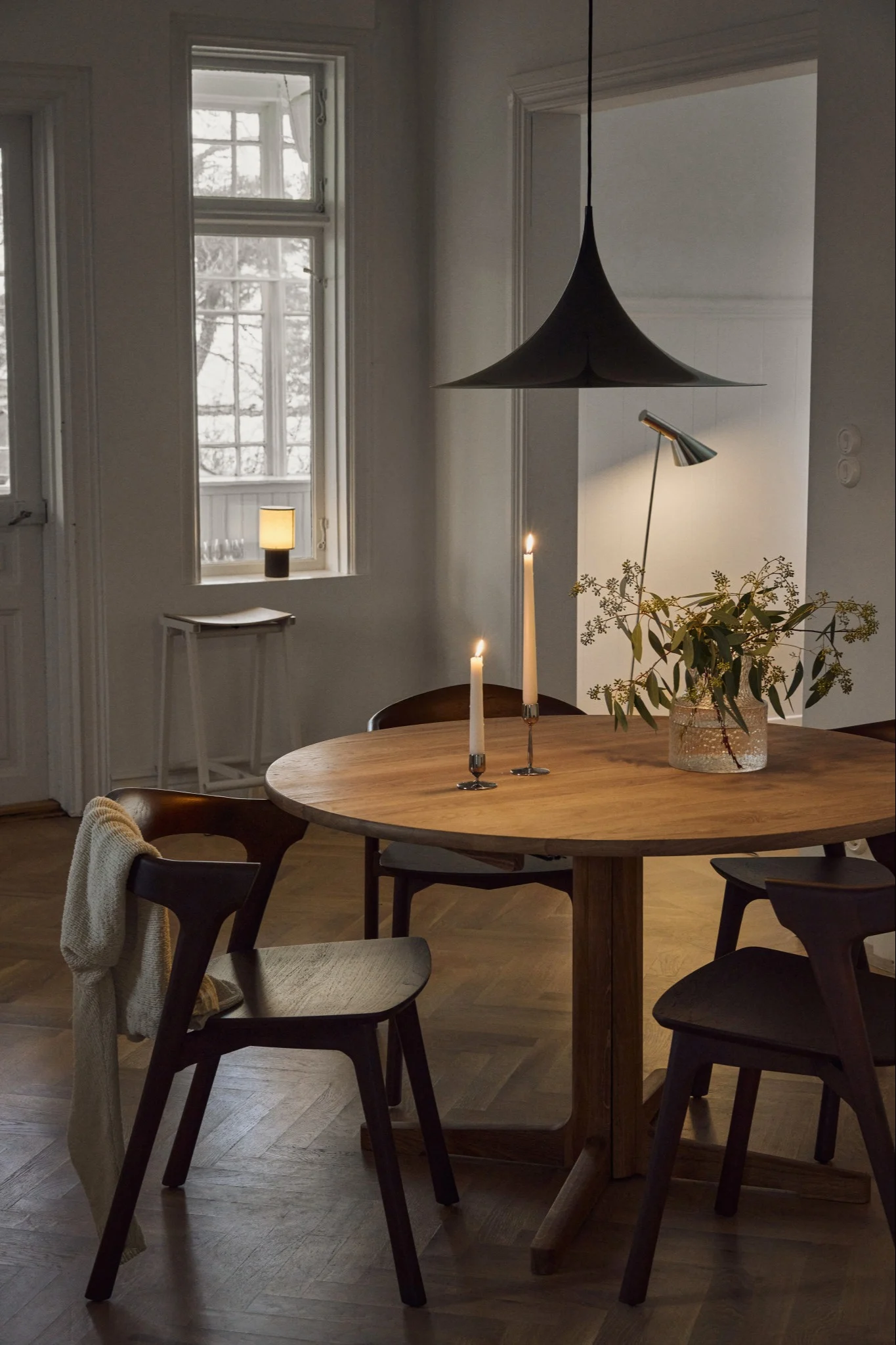 Mysig matsal med runt träbord, tända ljus, vas med grönska, mörka stolar och klockformad taklampa.