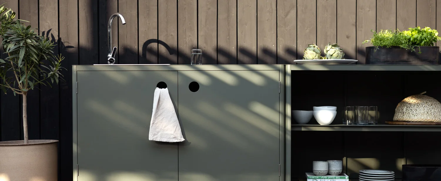 Utomhuskök med salviagröna skåp, diskho, örter och en krukväxt mot en mörk trävägg.