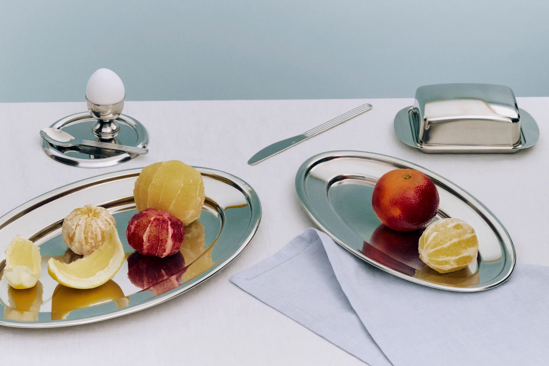 Frukost med skalade och oskalade citrusfrukter på silverbrickor, ett ägg i äggkopp och en smörask.