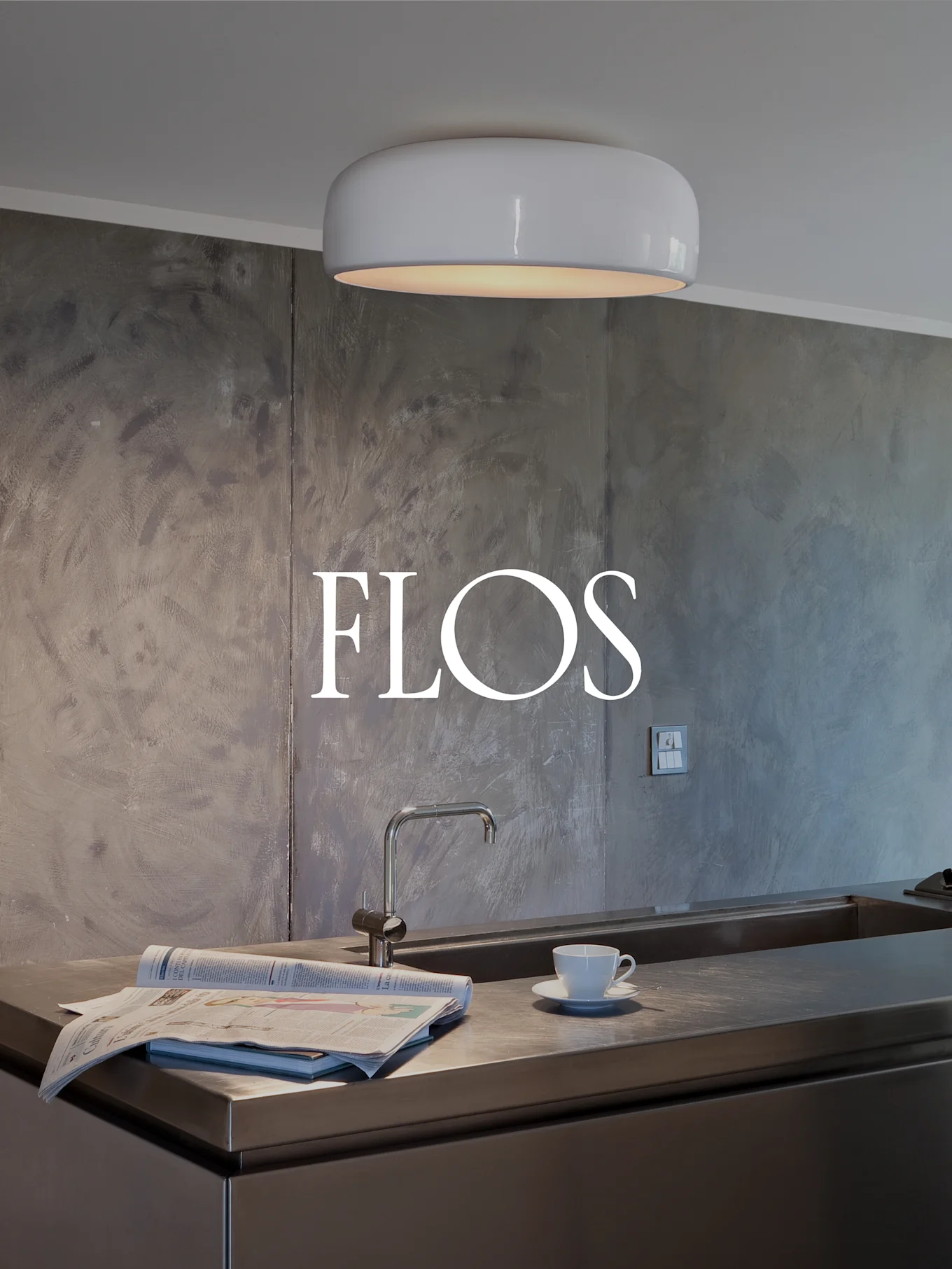 Modernt kök med vit, rund taklampa, bänkskiva i rostfritt stål med diskho, tidning och tekopp. FLOS-logotyp på texturerad vägg.