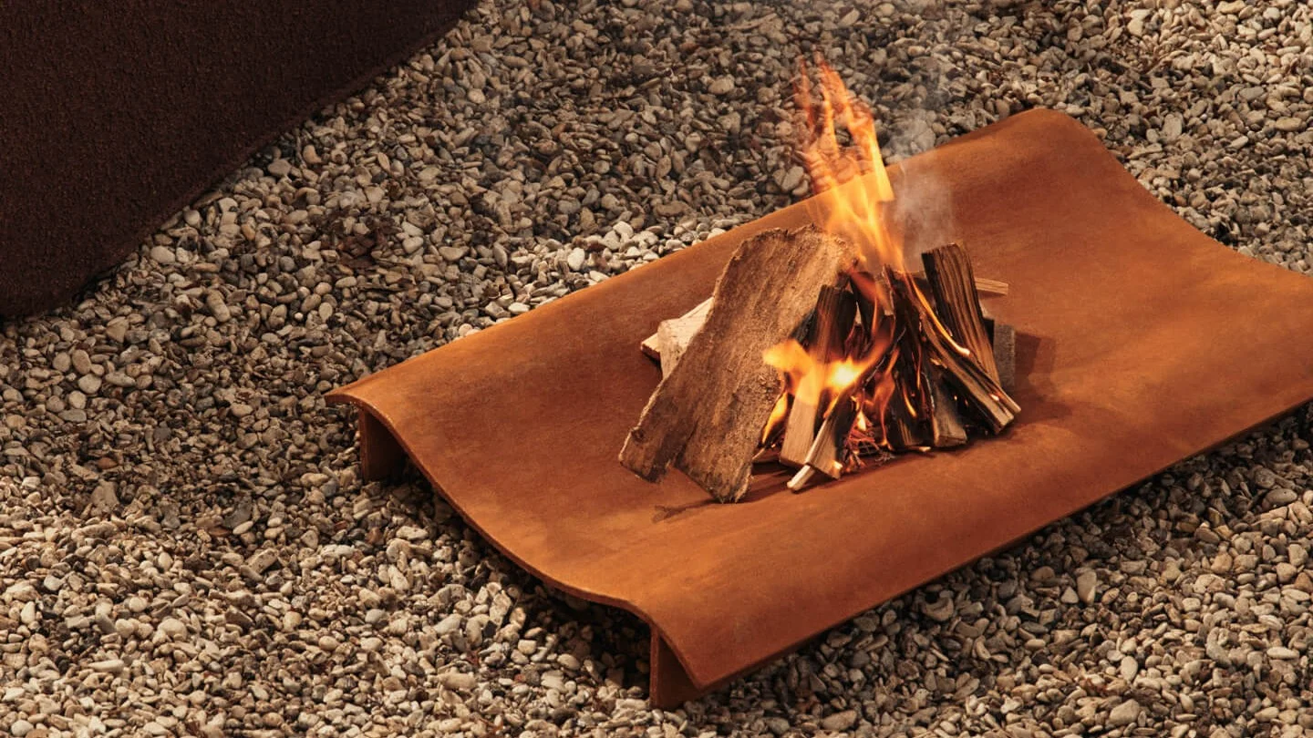 Liten brasa med brinnande ved på en rektangulär, rostig metallbricka över grus.