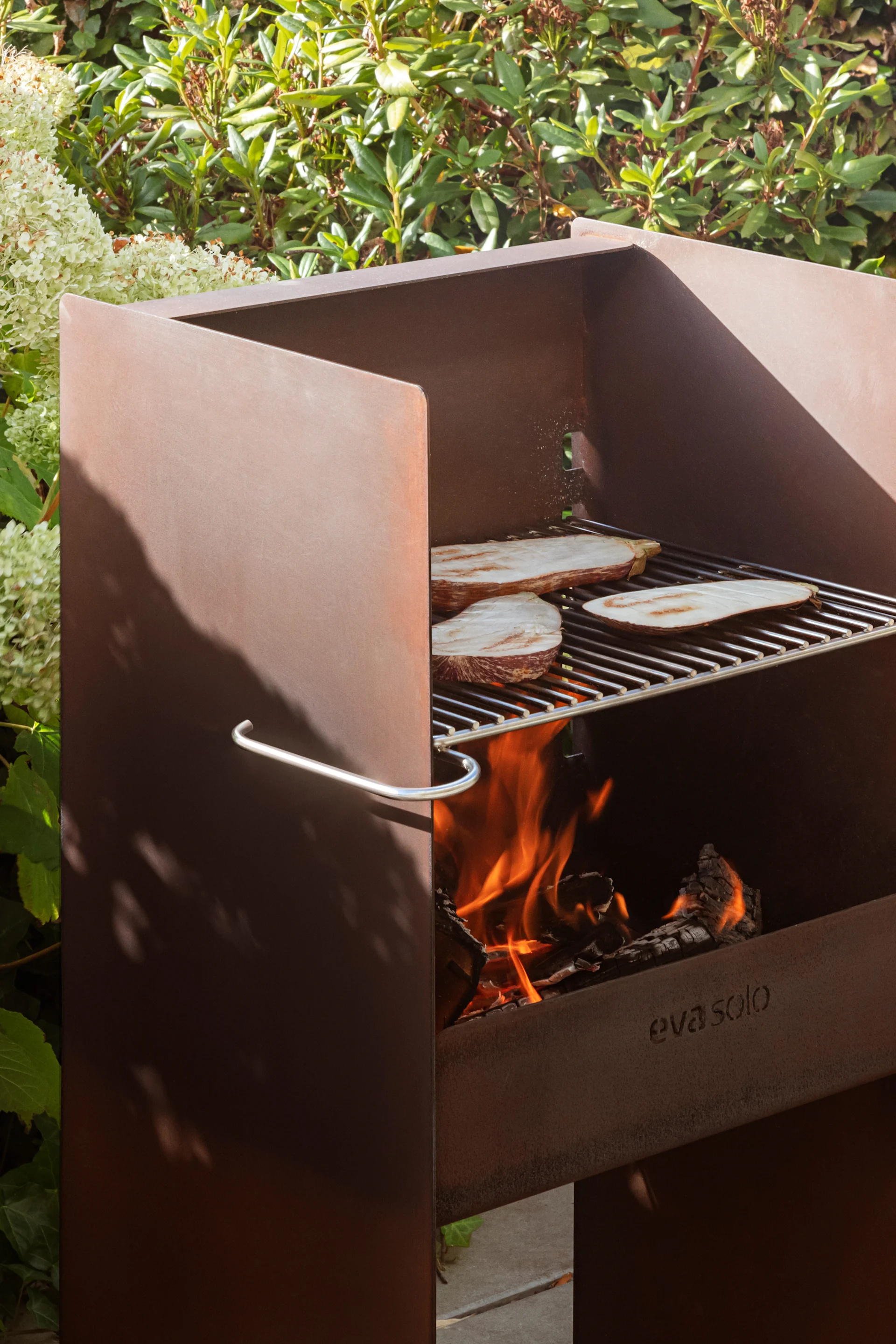 Utomhusgrill i cortenstål med aubergineskivor som grillas på galler över öppen eld, omgiven av gröna växter.
