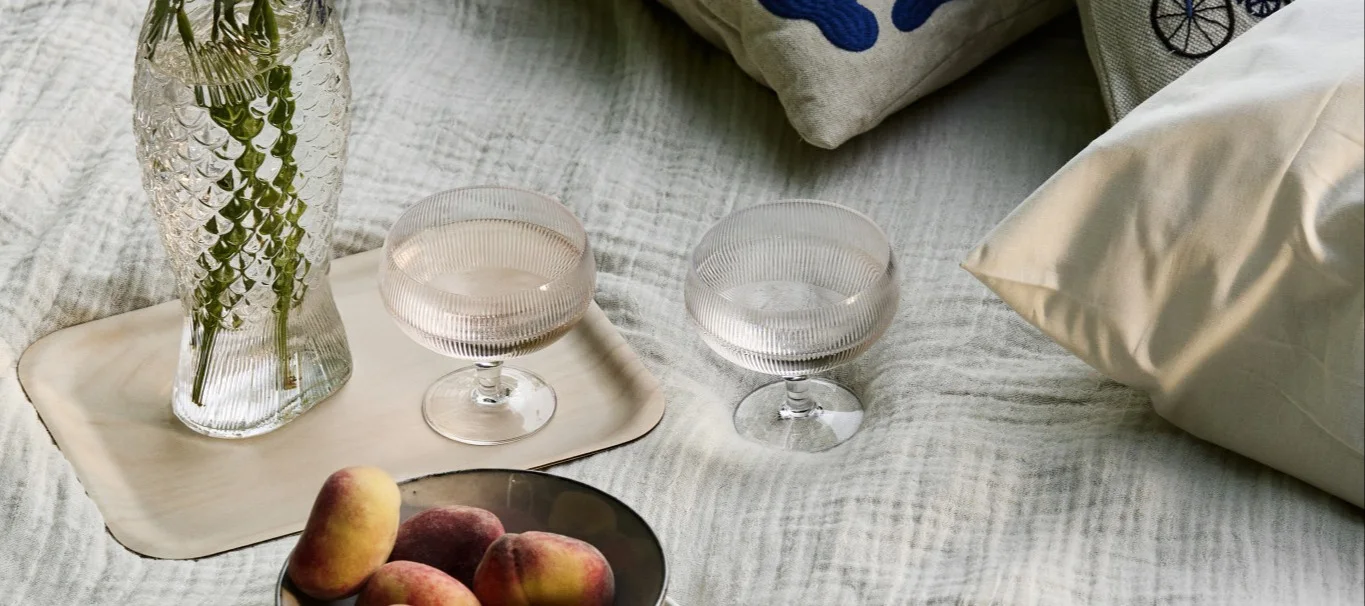 Picknickarrangemang med fiskformad vas, två räfflade glas och persikor på en ljus filt.