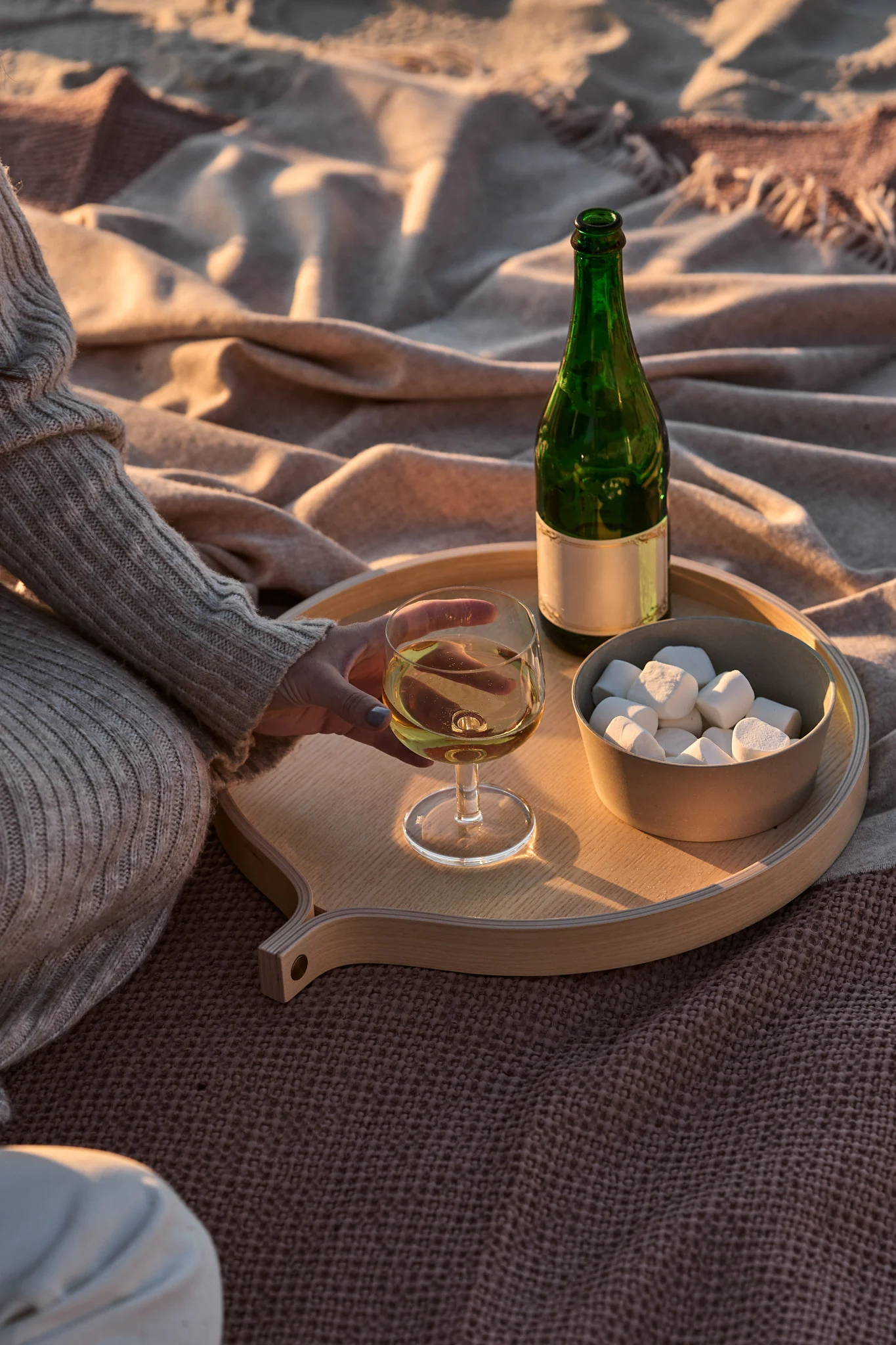 Person håller vitt vin på träbricka med grön flaska och marshmallows på filtar under den gyllene timmen.