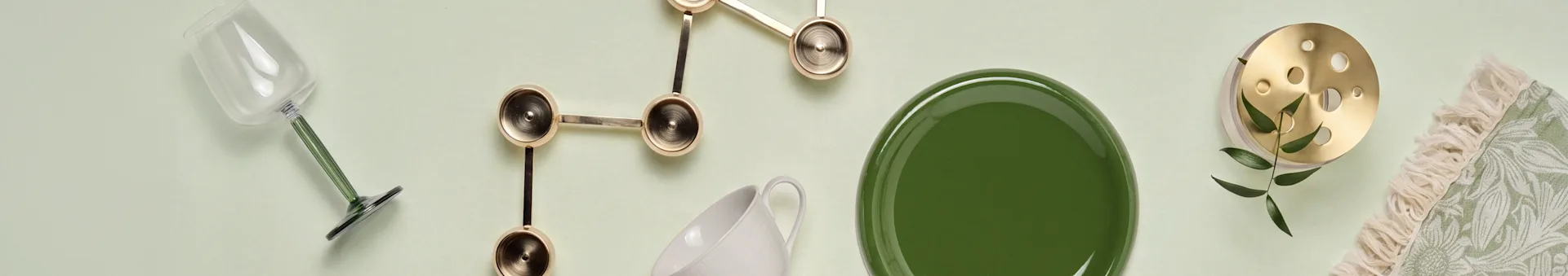 Glas med grön stam, gyllene skulptural inredning, vit mugg, grön tallrik, mässingsföremål med blad och grön fransad textil.