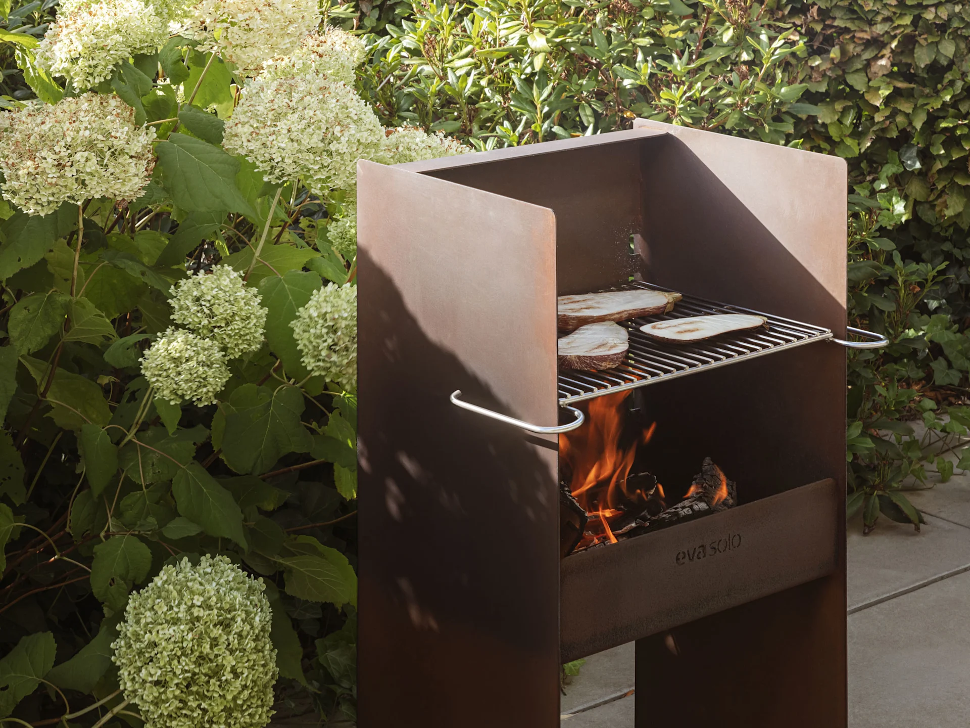 En rustik utomhusgrill i metall med lågor och aubergineskivor som grillas på gallret.