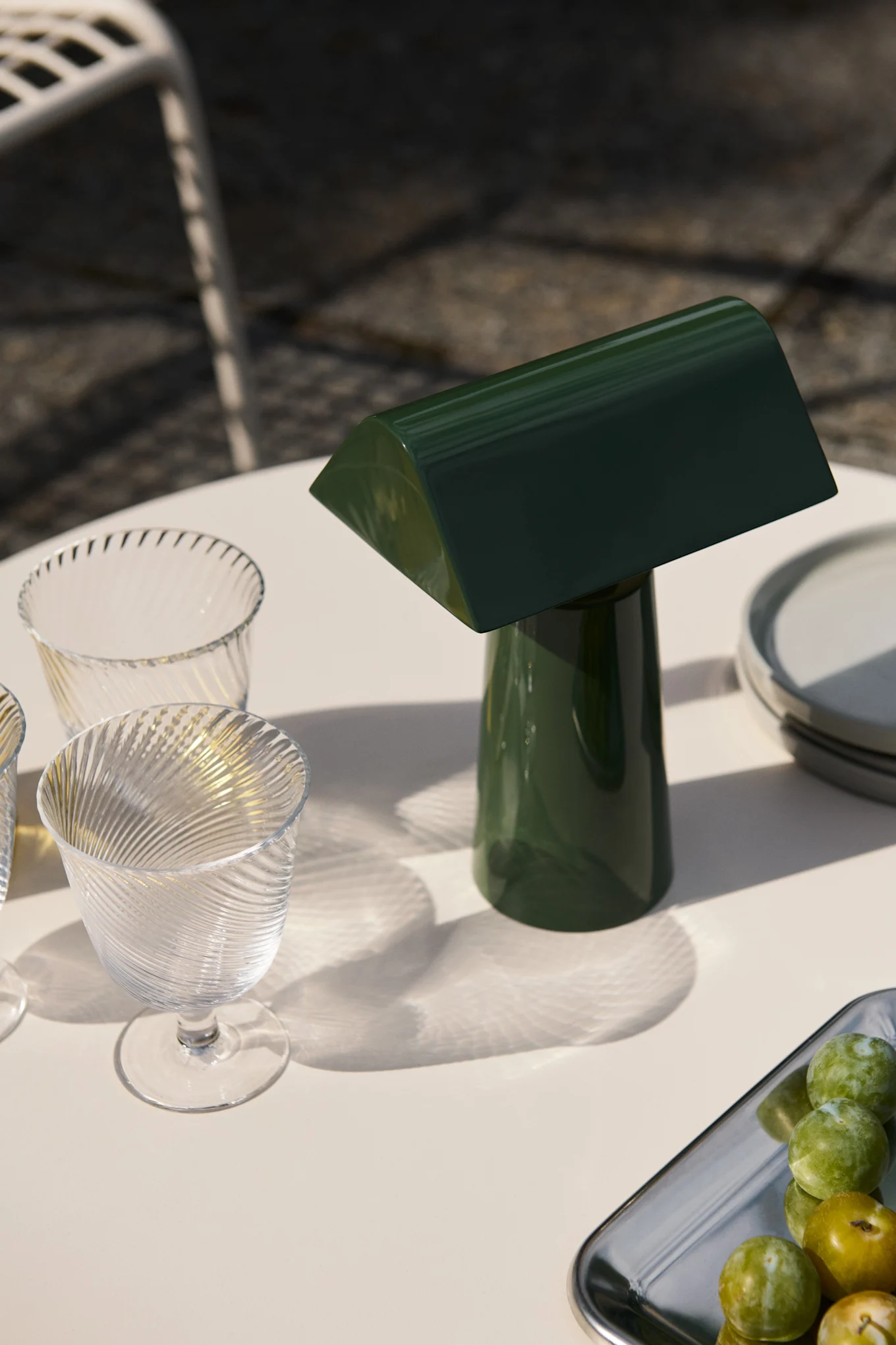 En mörkgrön bordslampa och två räfflade glas på ett utomhusbord med plommon på en bricka, badade i solljus. Inspiration till din balkong 2026.