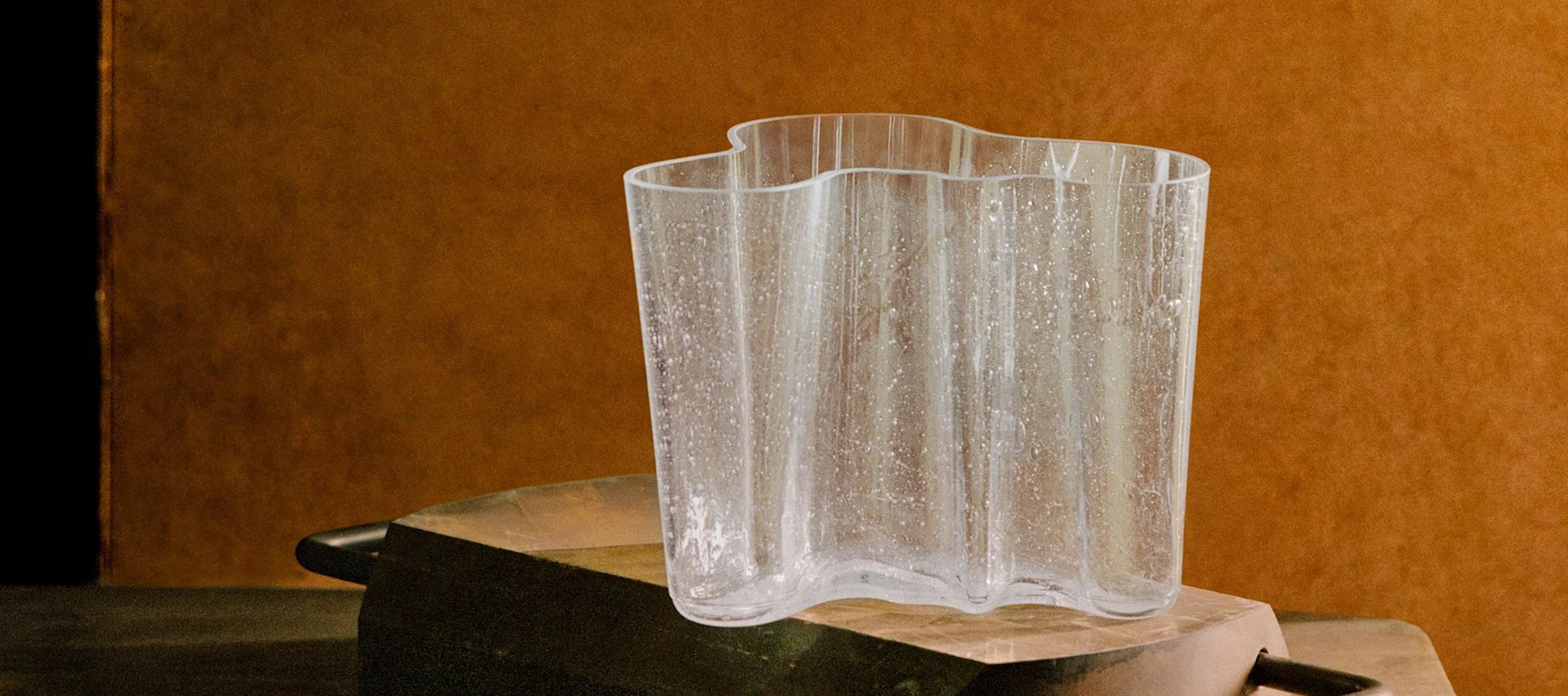 En klar, vågig glasvas med texturerade fläckar på en rustik mörk yta mot en varmbrun bakgrund.