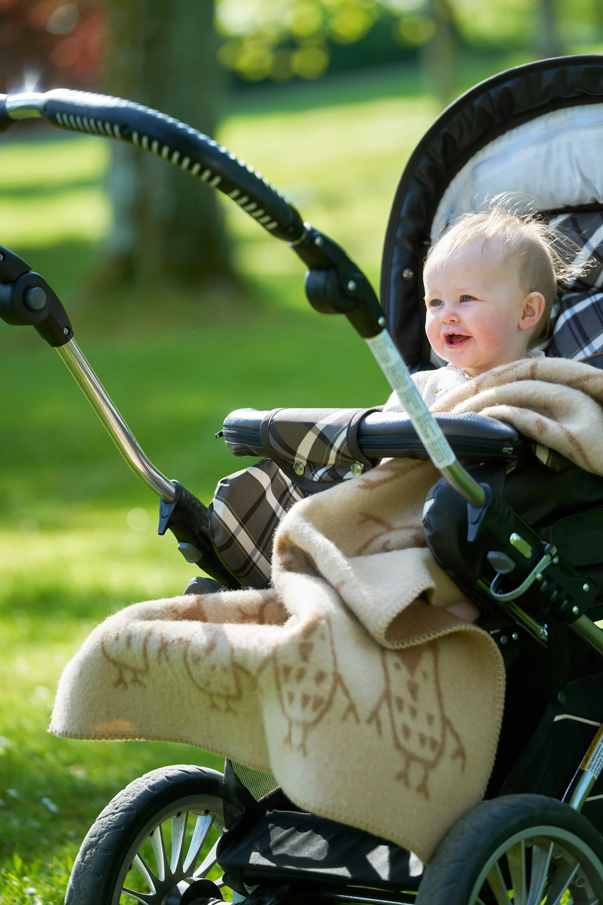 En leende bebis med ljust hår, inlindad i en filt med ugglemönster, sitter i en barnvagn utomhus i en solig park.