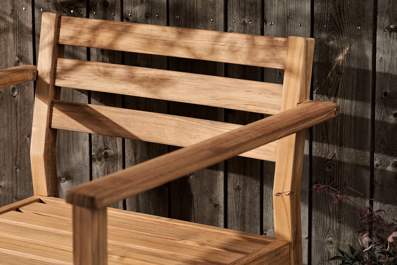 Lamellträstol med armstöd mot ett mörkt trästaket, täckt av bladskuggor.