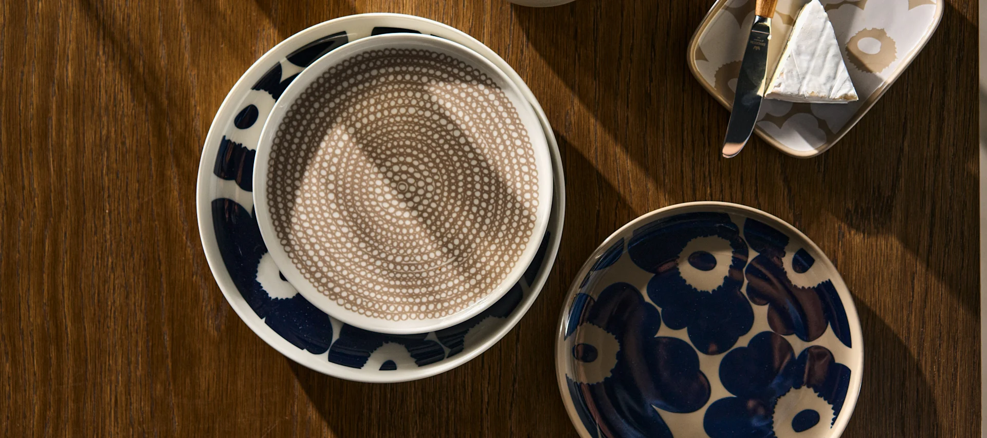 På ett träbord ligger Marimekkos Unikko tallrik i färgen vit-mörkblå, Unikko assiett i färgen terra-mörkblå, Unikko fat i beige och så en Siirtolapuutarha tallrik i färgen white-clay. 