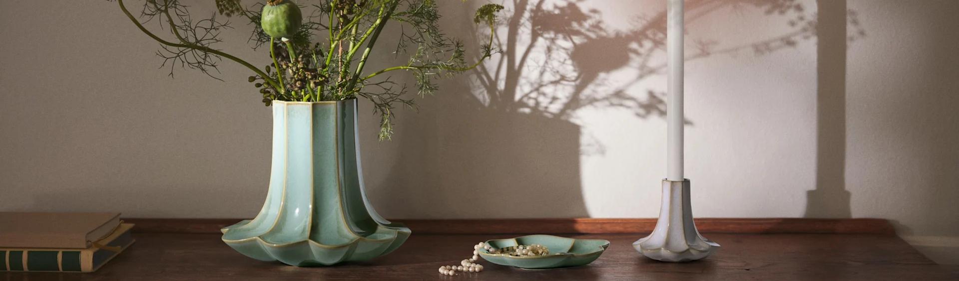 En räfflad turkos vas med bladverk, ett pärlhalsband i en matchande skål, ett vitt ljus i en räfflad hållare och böcker på en träyta.