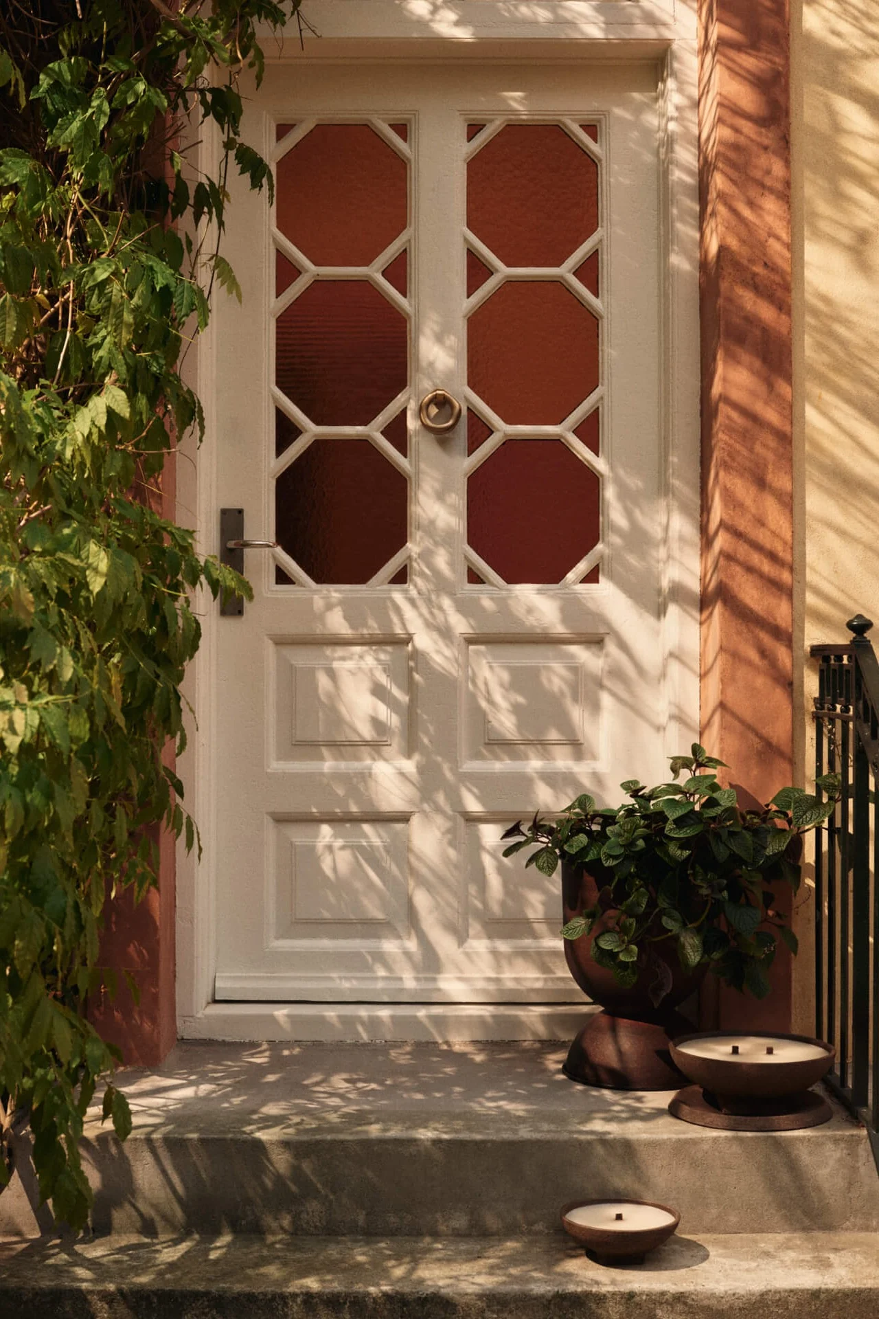 Vit ytterdörr med åttakantiga glaspaneler, gröna klätterväxter till vänster och en kruka med två ljus på trappan.