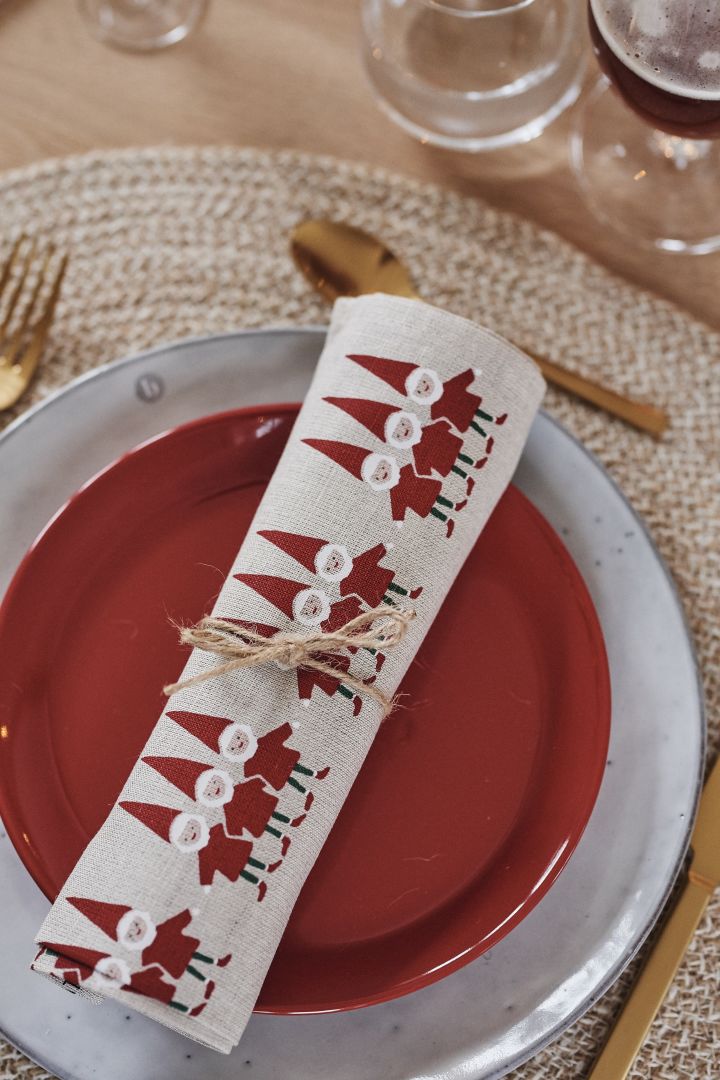 Ihoprullad kökshandduk med tomtemotiv vilar på röd assiett och Nordic Sand tallrik som blir tallrikskuvertet för en röd juldukning. 