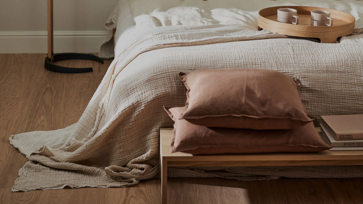 Säng med ljust täcke, två roströda kuddar på ett nattduksbord och en träbricka med två koppar på sängen.