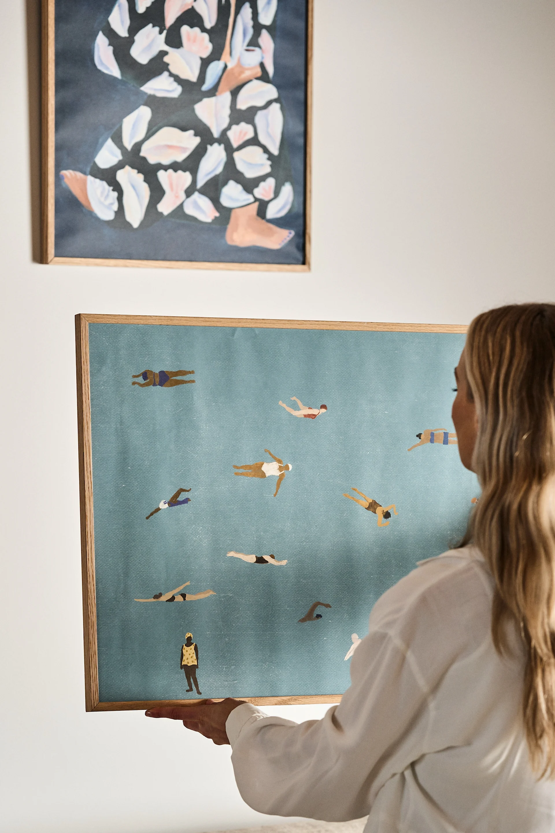 En kvinna håller upp Swimmers poster från Fine Little day mot en naturvit vägg. Postern har motiv föreställer ett flertal människor som simmar i ett hav. 