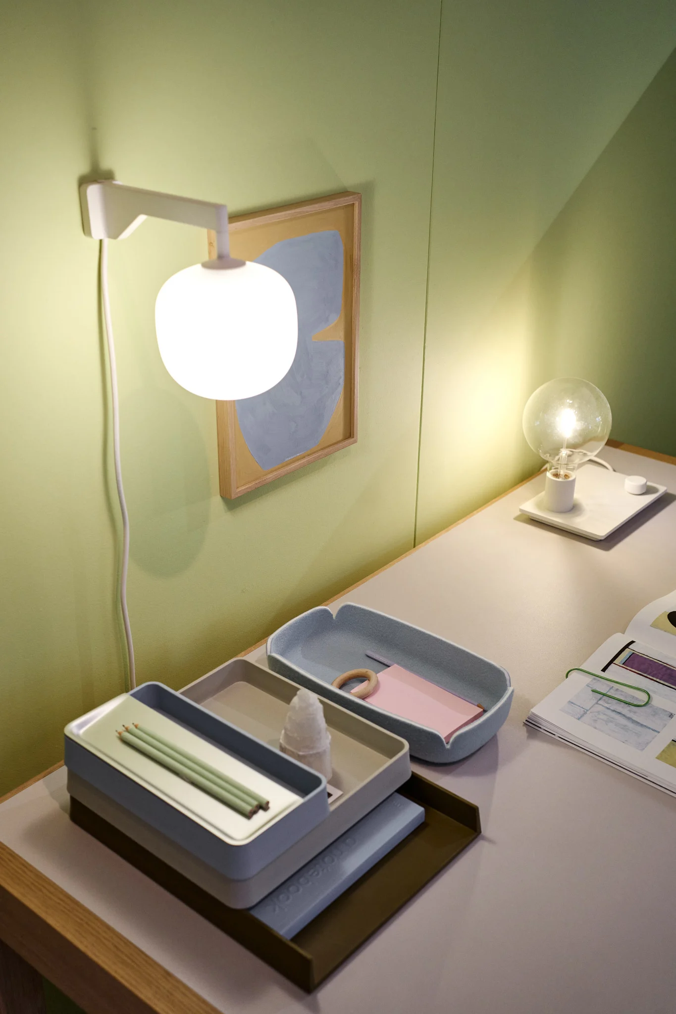 Ett prydligt skrivbord framför en grön vägg med den beigefärgade vägglampan Rime från Muuto.