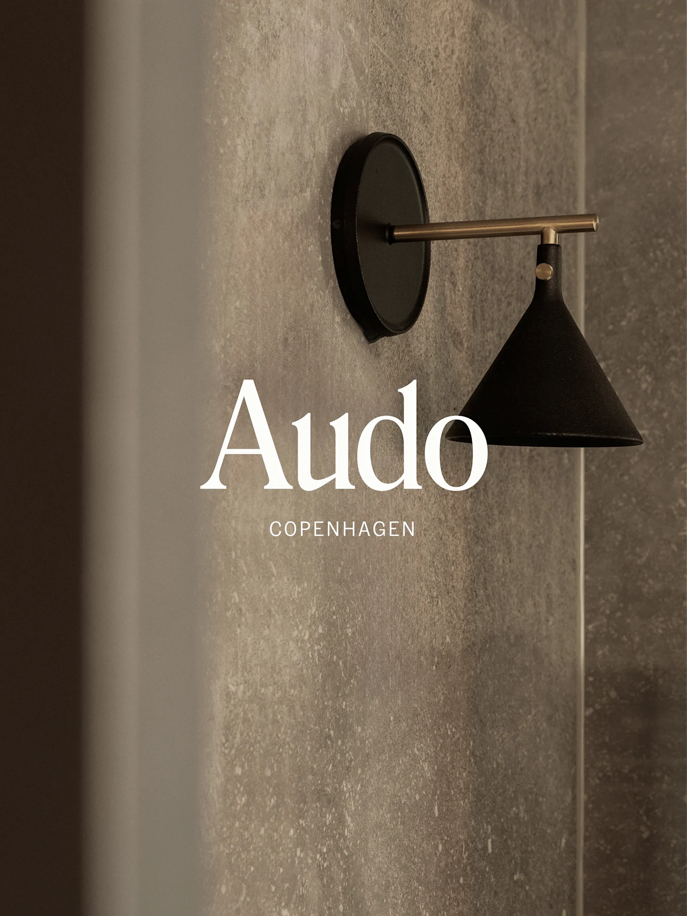 En väggmonterad svart lampa med mässingsarm mot en texturerad grå vägg, med texten 'Audo Copenhagen'.