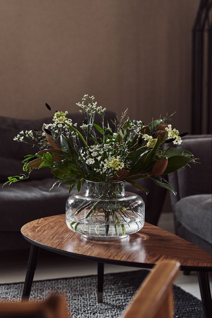 Urna vas i färgen clear från varumärket Marimekko står på ett soffbord i valnötsfärgat trä, innehållandes en bukett med gröna blomster. 