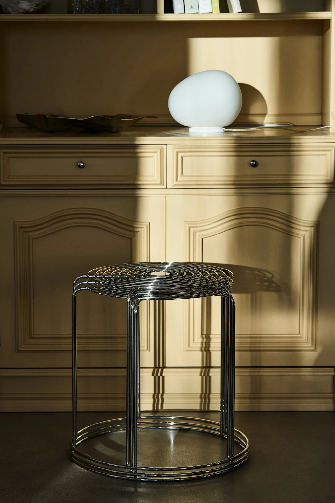 En stapel kromade metallpallar framför ett ljusbrunt skåp med en vit äggformad lampa, badat i solljus.