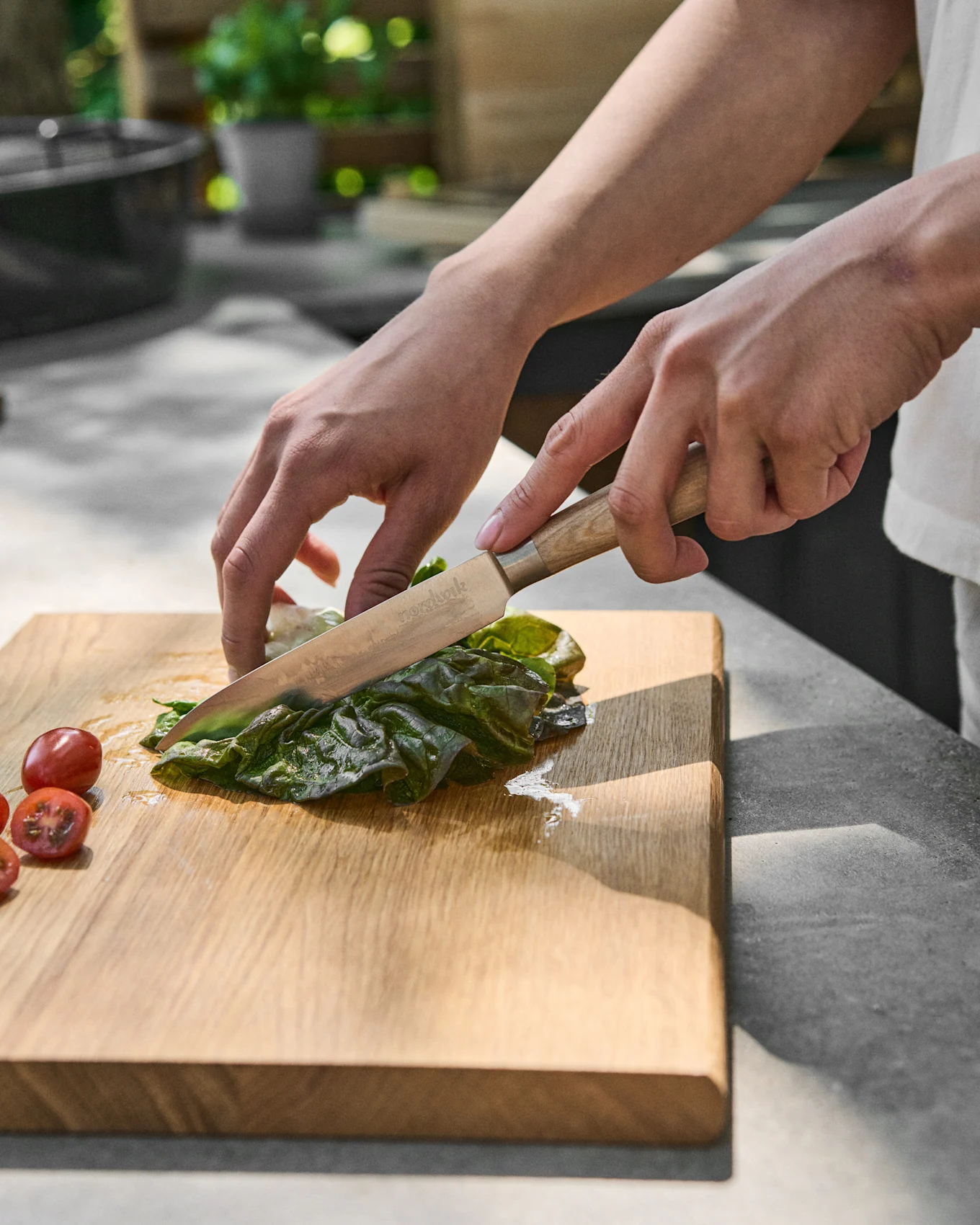Händer skivar grön sallad på en träskärbräda bredvid körsbärstomater.