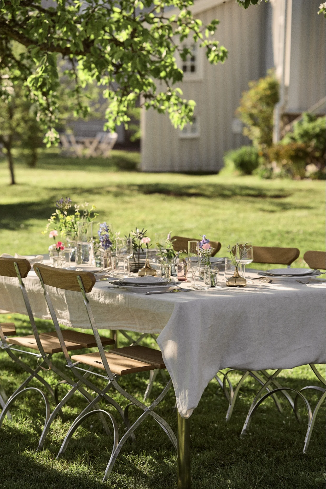 Ett utomhusmatbord dukat med vita tallrikar, klara glas och små blomsterbuketter på en linneduk i en solig trädgård.