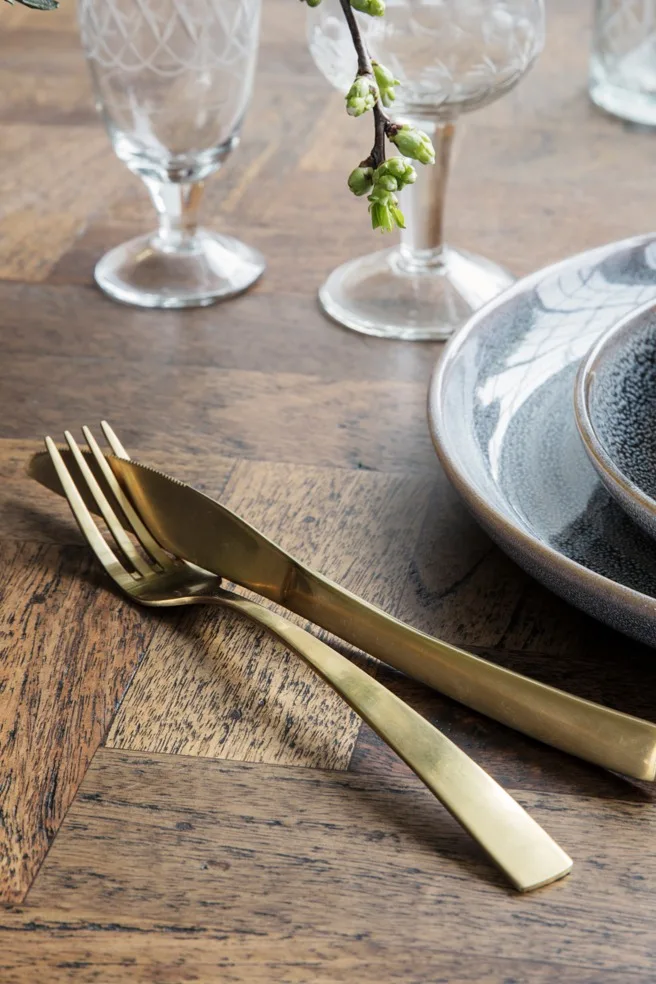 Guldfärgad gaffel och kniv på ett träbord bredvid keramiktallrikar och mönstrade glas, med en liten gren med gröna knoppar.