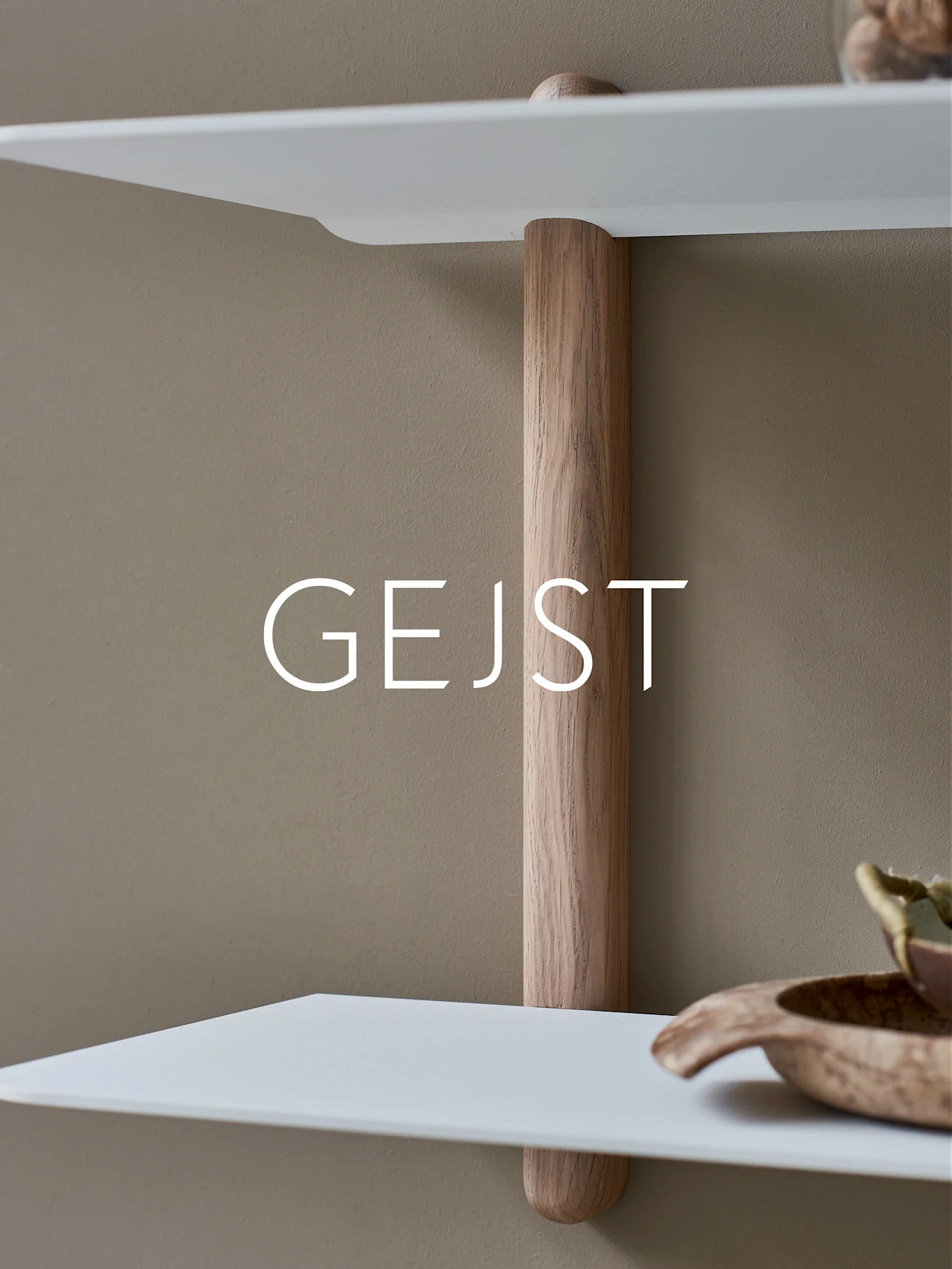 Två vita, rundade hyllor förbundna med en vertikal ljus trästödpåle mot en beige vägg. En träskål står på den nedre hyllan.