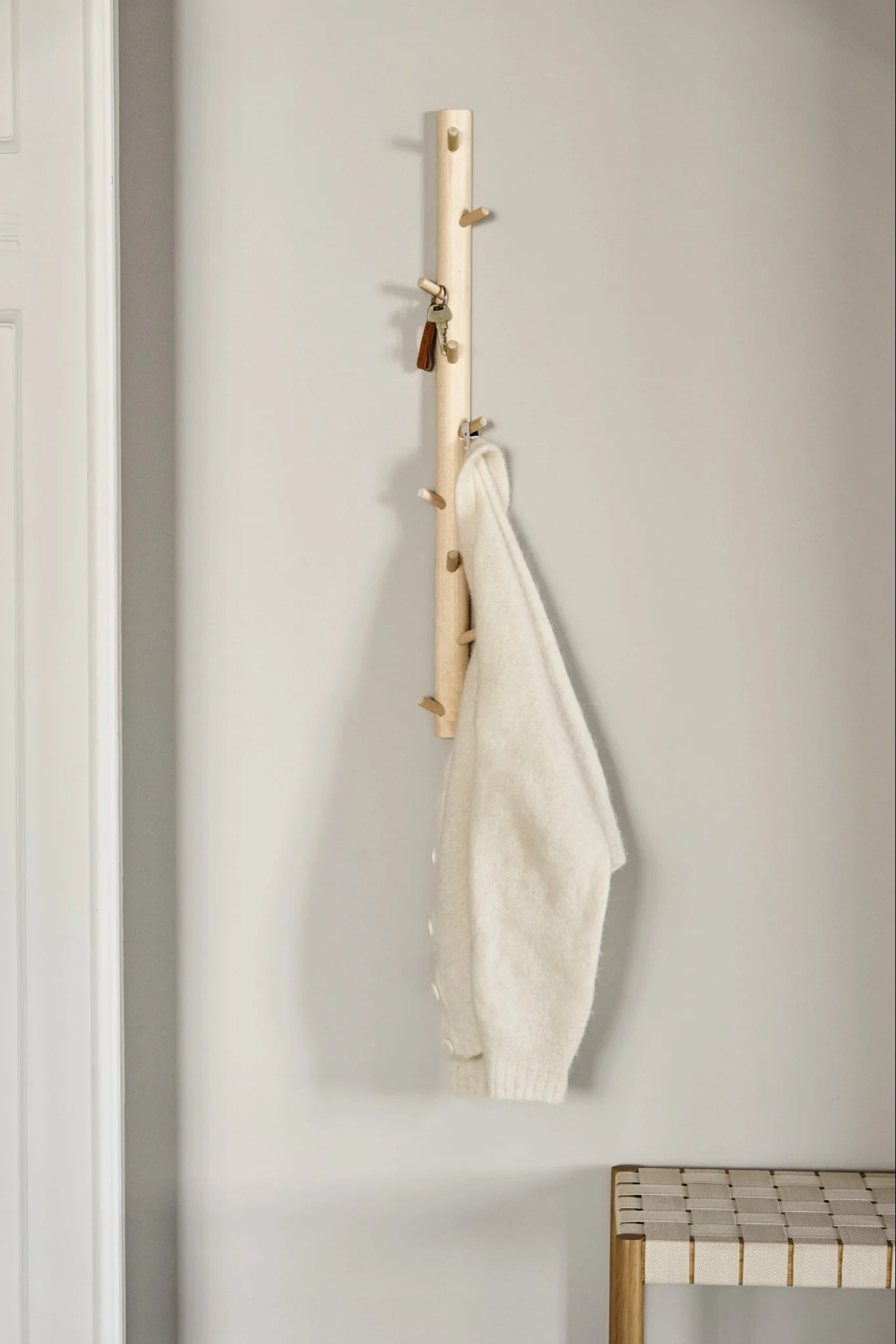 En ljus träkroklist med krokar håller nycklar och en gräddvit tröja, med en flätad bänk under.