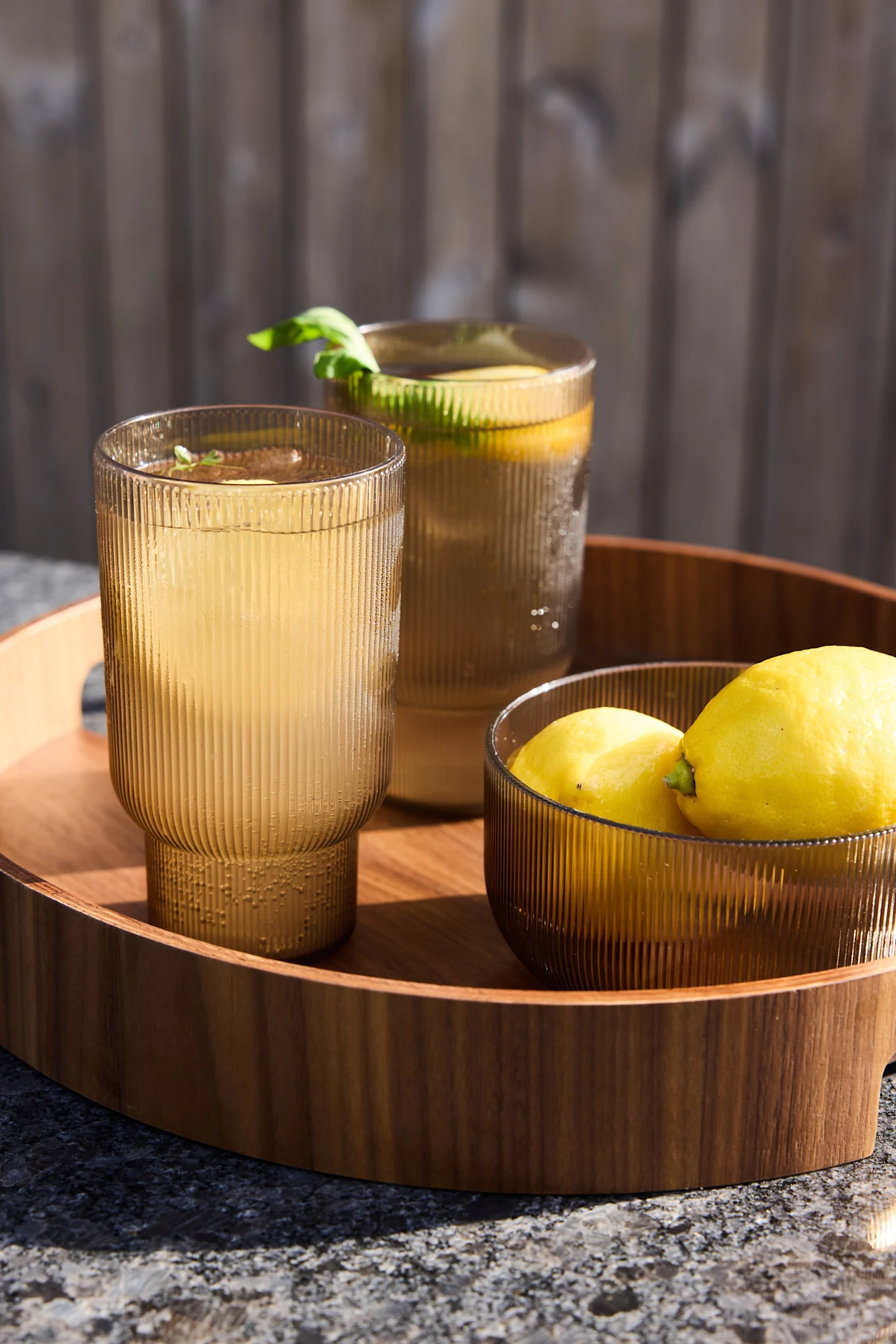 Två räfflade bärnstensfärgade glas med lemonad och en skål citroner på en träbricka utomhus.
