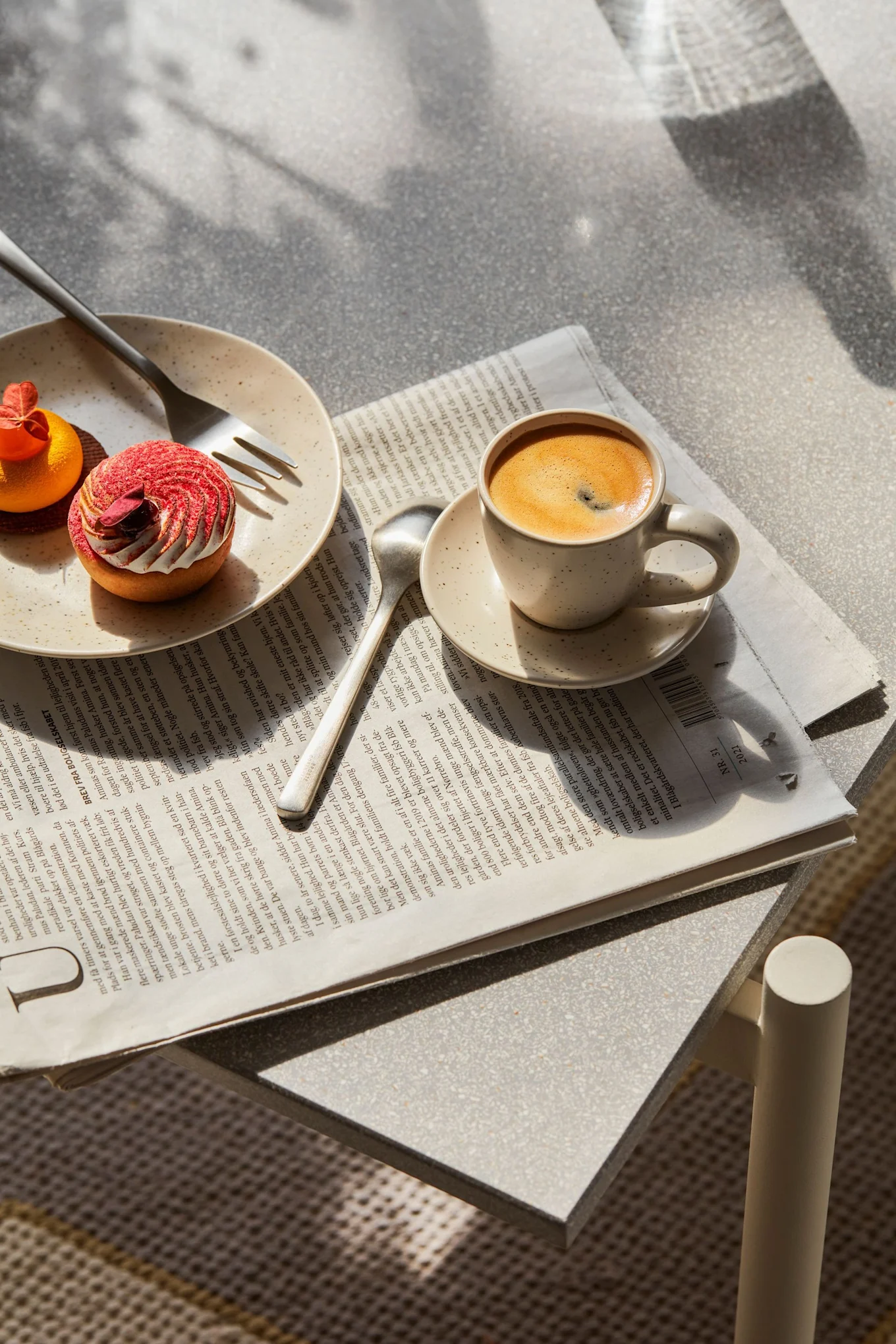Kaffekopp, sked och tallrik med två bakverk (rött och orange) på en tidning, på ett spräckligt bord med fläckiga skuggor.