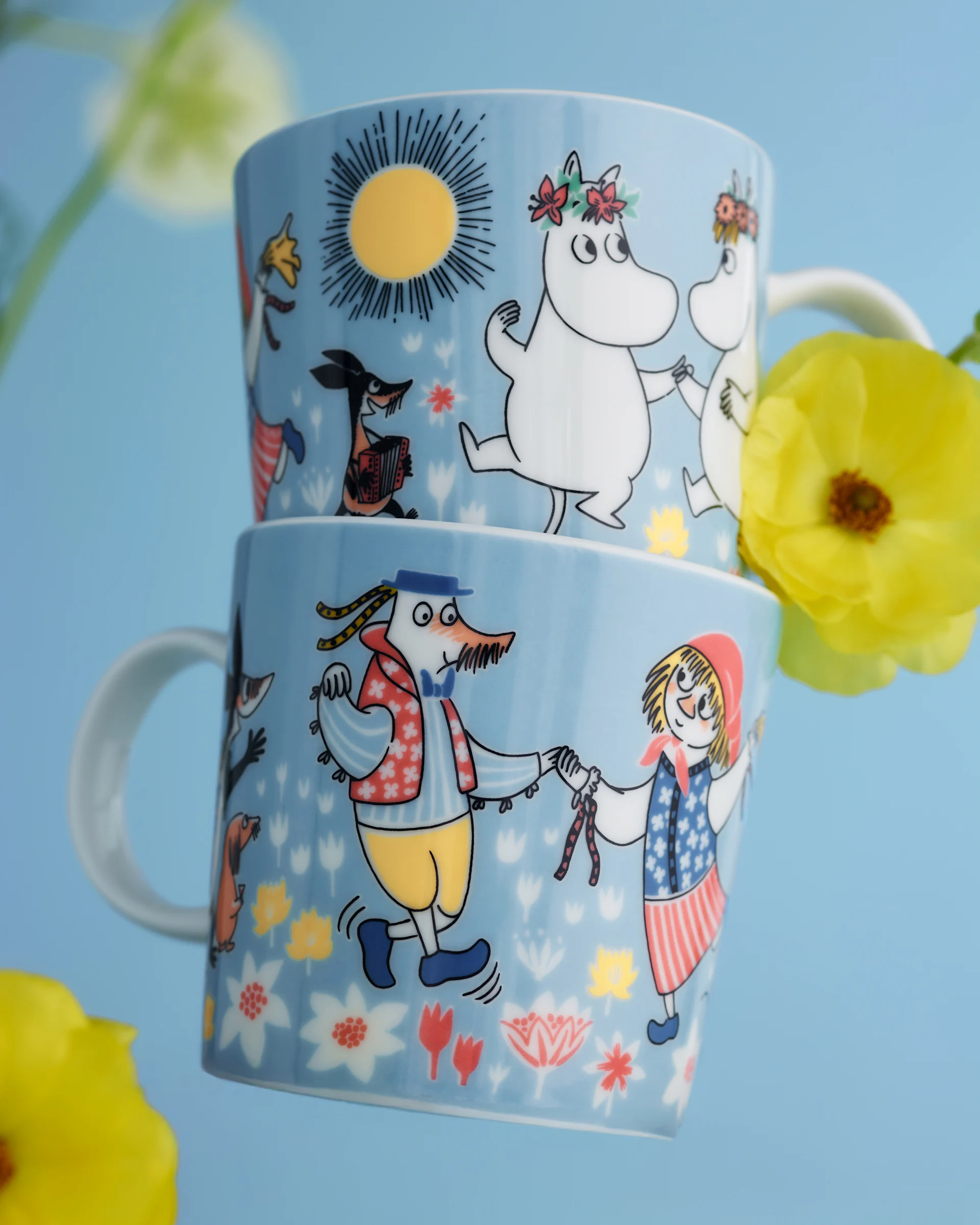 Två ljusblå Muminmuggar, staplade, med dansande och musikspelande karaktärer bland blommor, med gula ranunklar.