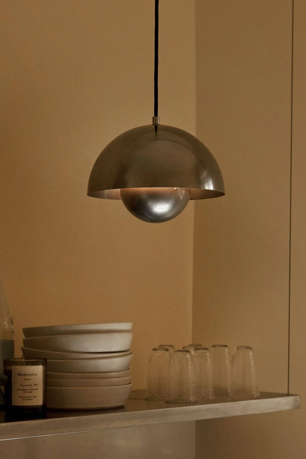 En kupolformad metallpendellampa hänger över en hylla med skålar, glas, flaskor och ett ljus mot en varmbrun vägg.