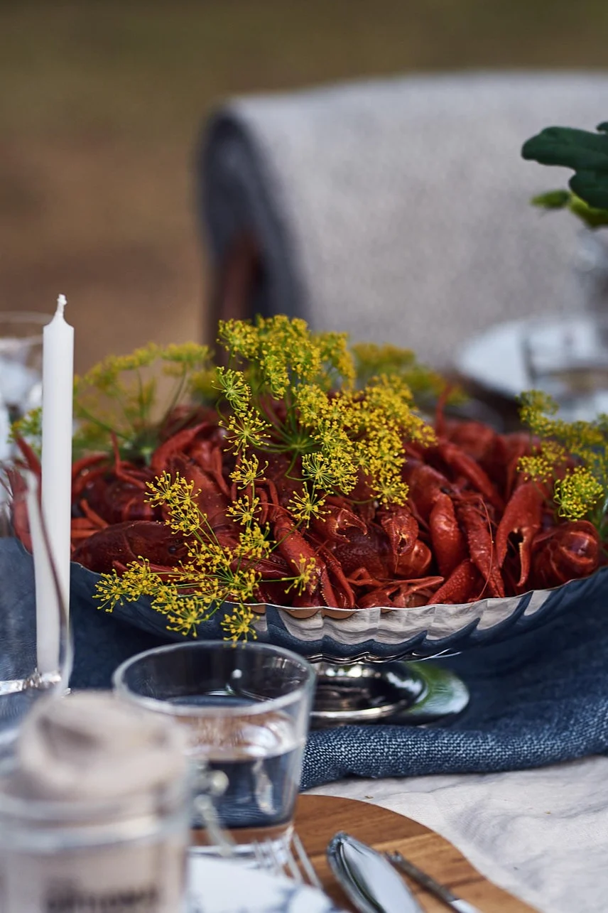 Utomhusbord dukat med en silverbunke med röda kräftor och dill, levande ljus och ett blomsterarrangemang.