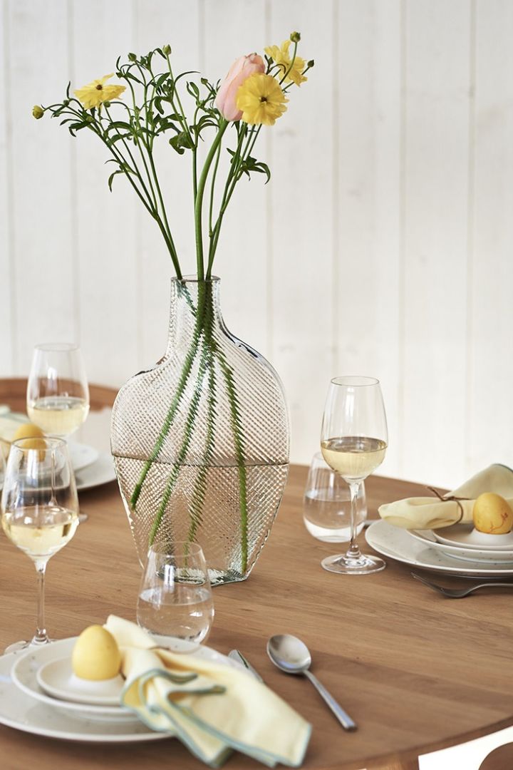 En gul påskdukning med vas i klarglas på bordet och med gula vårblommor i. 