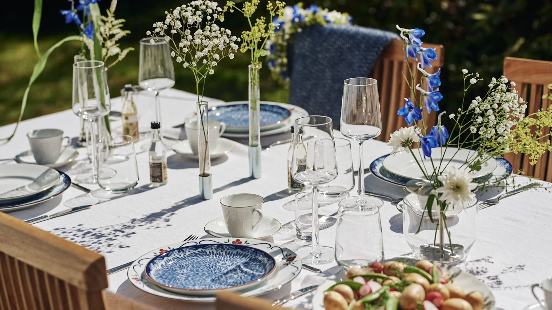Elegant utomhusbord dukat med vit duk, blåmönstrade tallrikar, vinglas, koppar och vaser med blommor.
