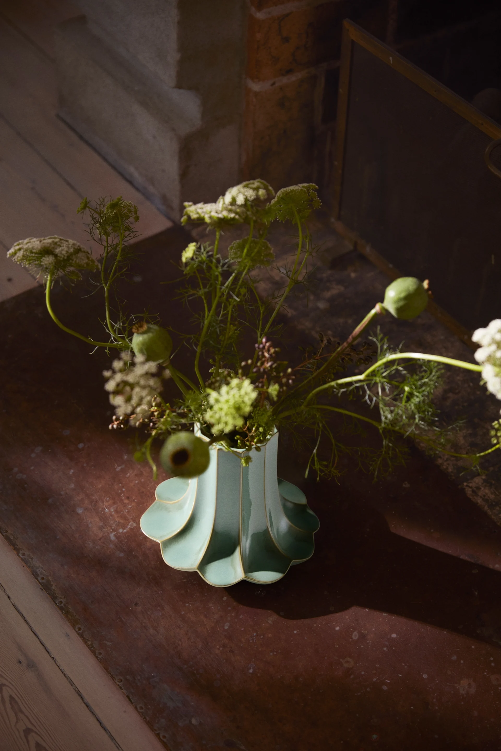 En ljus turkosgrön vågig vas med vita blommor, gröna blad och vallmofrökapslar, på en mörk yta nära spis.
