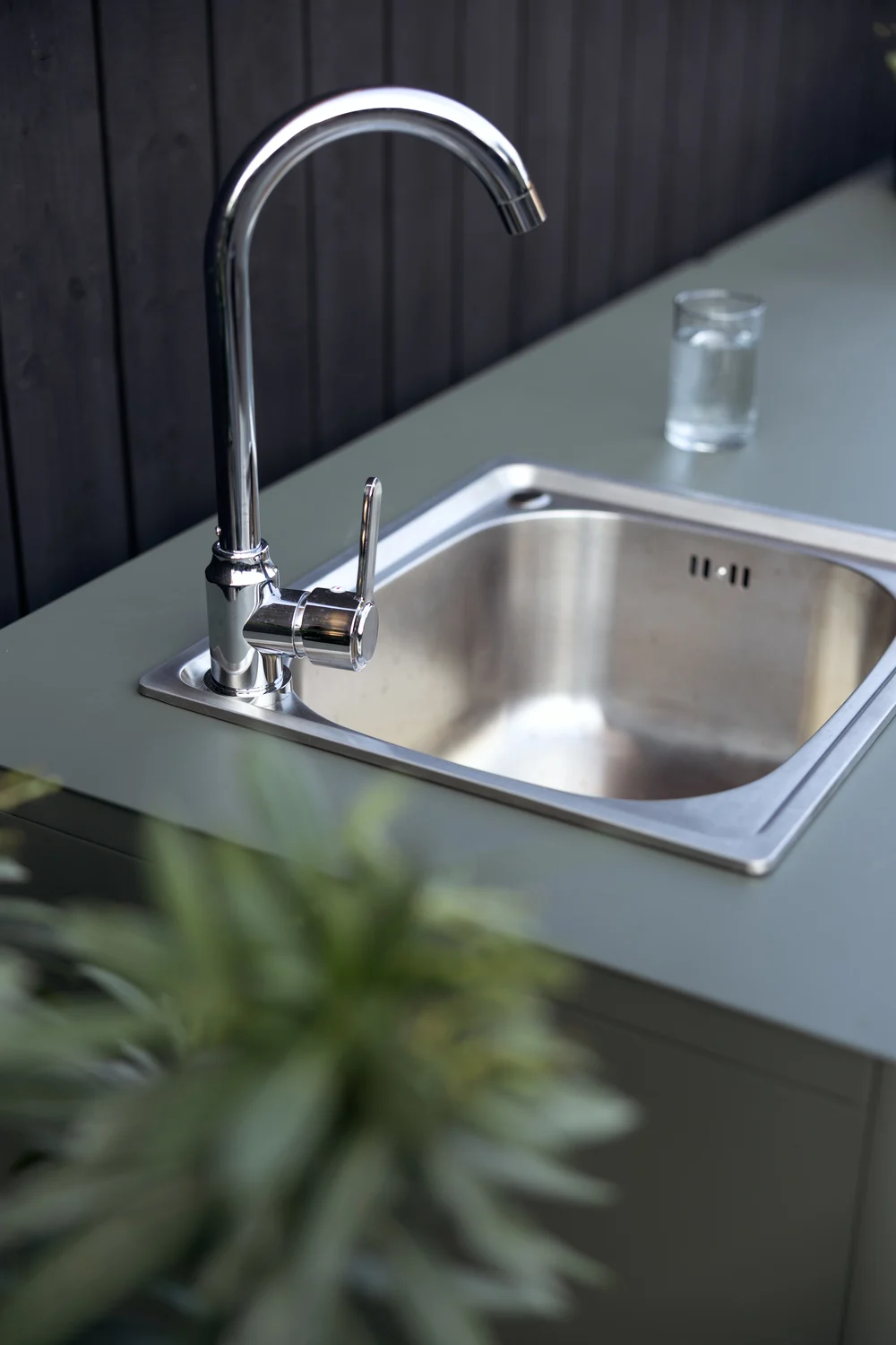 Utomhusköksvask med kromkran och ett glas vatten på en grön bänkskiva.